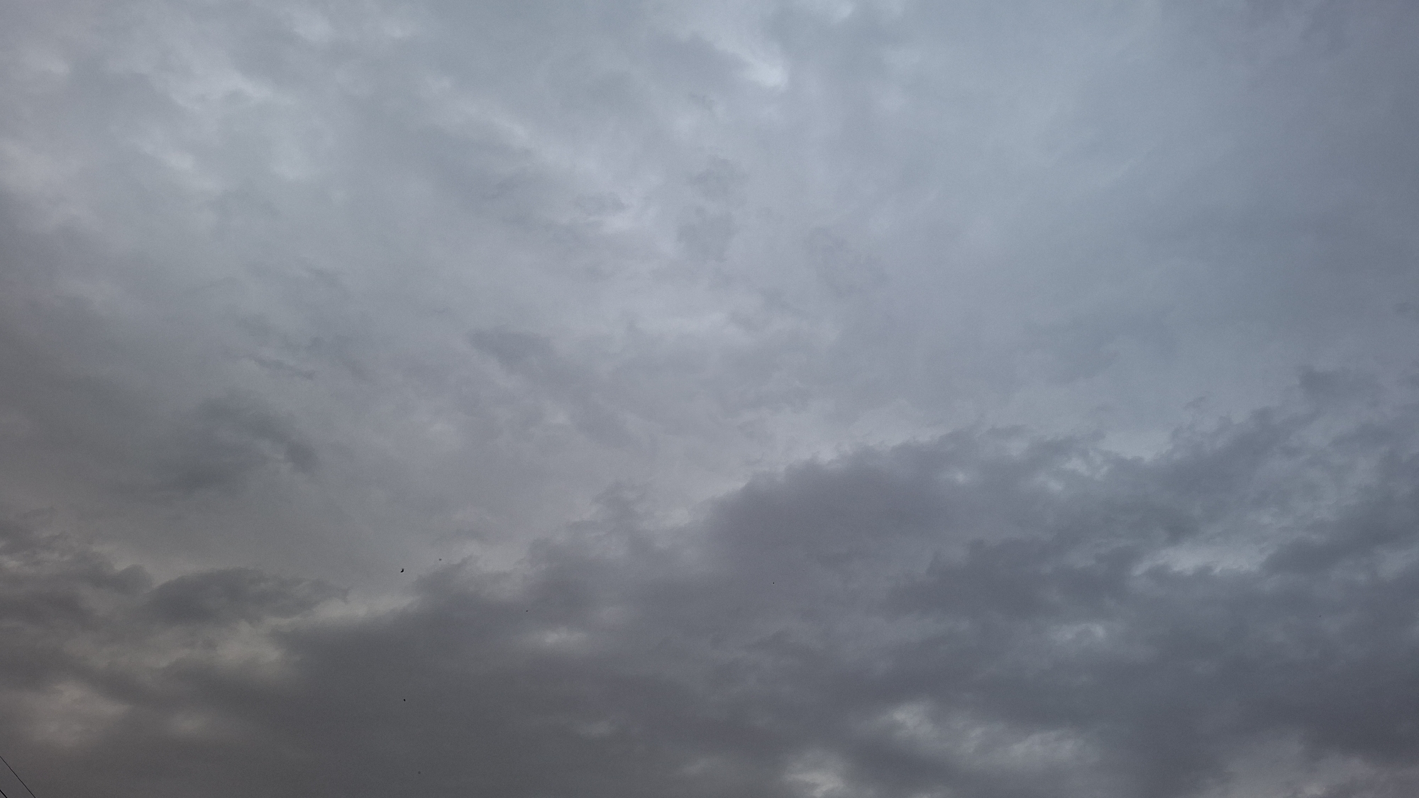 The image depicts an overcast sky with dense, dark clouds dominating the lower portion of the frame. The upper part of the sky is lighter, with softer, diffused clouds. There are a few small, dark shapes that may be birds or debris visible in the sky, particularly in the lower left and middle right sections. The overall impression is of a grey, somber sky, possibly indicative of impending rain or a storm. There are no visible structures or geographical features of Shyriaieve, Ukraine.