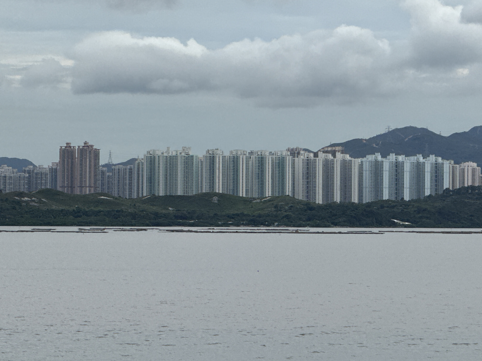 The image depicts a wide, calm body of water in the foreground, with a dense line of high-rise apartment buildings stretching across the horizon behind a strip of lush green hills. The sky is overcast with thick, gray clouds. The buildings are numerous, appearing to form a continuous urban landscape. Some smaller structures and darker hills can be seen in the background, further inland. In the water, there are several long, rectangular floating structures, possibly for aquaculture or some form of water management. The scene suggests a coastal or riverside urban environment, characteristic of densely populated areas. The location is identified as Futian District, China.