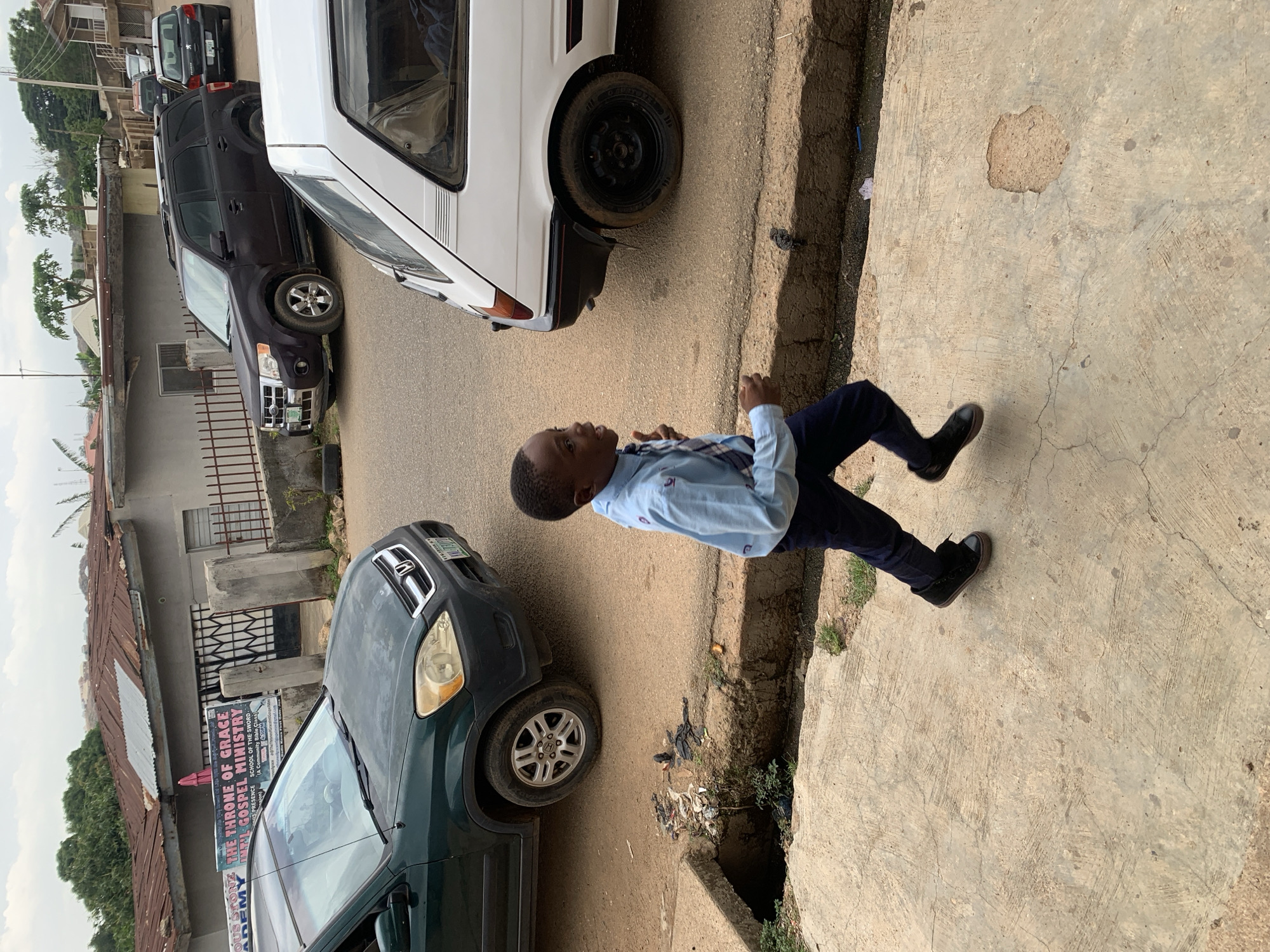 In Jos, Nigeria, a young boy in a light blue collared shirt and dark pants runs along a cracked concrete sidewalk next to a drainage ditch filled with debris. A dark green SUV is parked on the street to the left, partially obscuring a sign for "THE THRONE OF GRACE GOSPEL MINISTRY". To the right, a white hatchback car is also parked on the street. The background shows a residential area with corrugated metal roofs and a hazy sky.