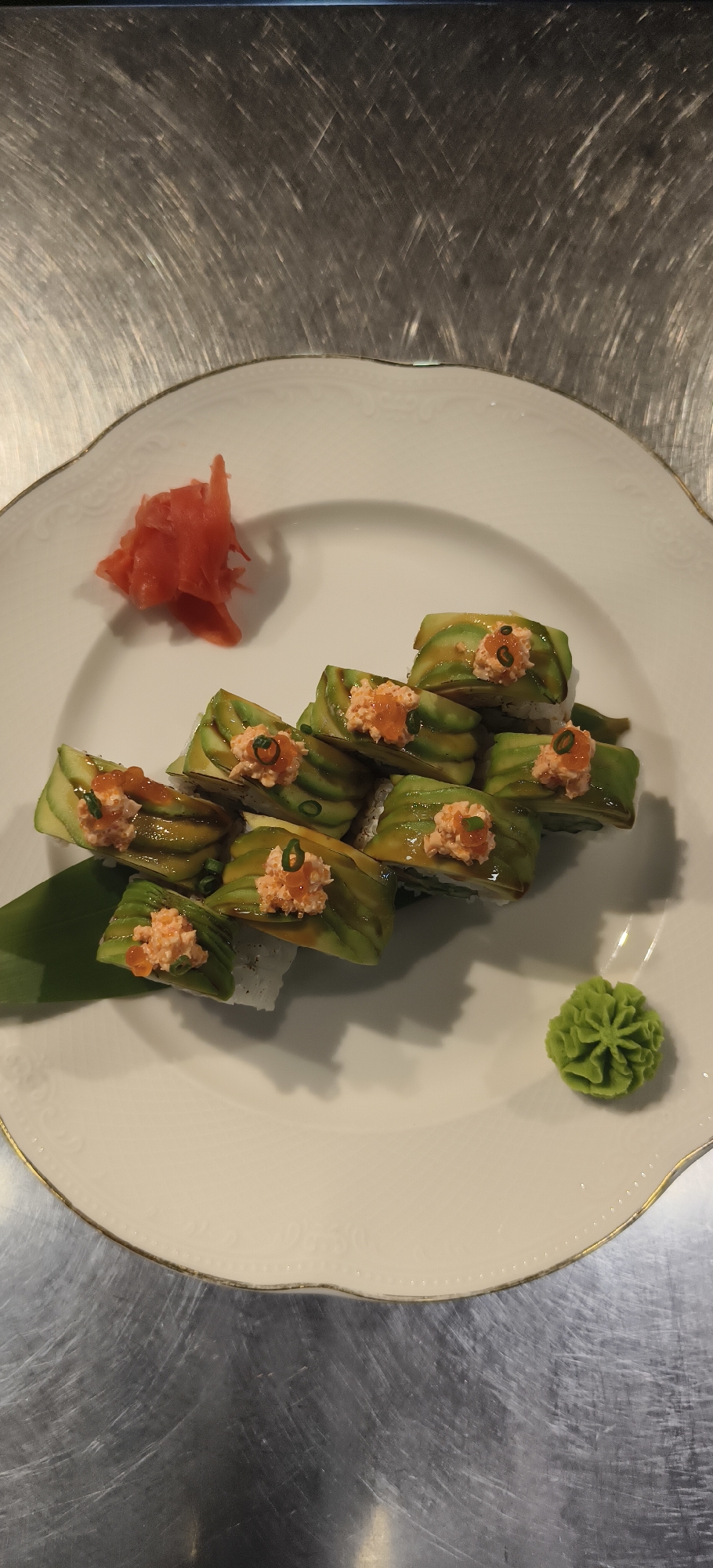 On a white plate with a gold trim, a roll of sushi sits arranged in a cascade, each piece topped with avocado, what appears to be salmon, and garnished with tobiko and scallions. Beside the sushi are condiments: a mound of pickled ginger and a dollop of wasabi. The plate is set against a stainless steel countertop. This scene is located in the city of Unknown, Unknown.
