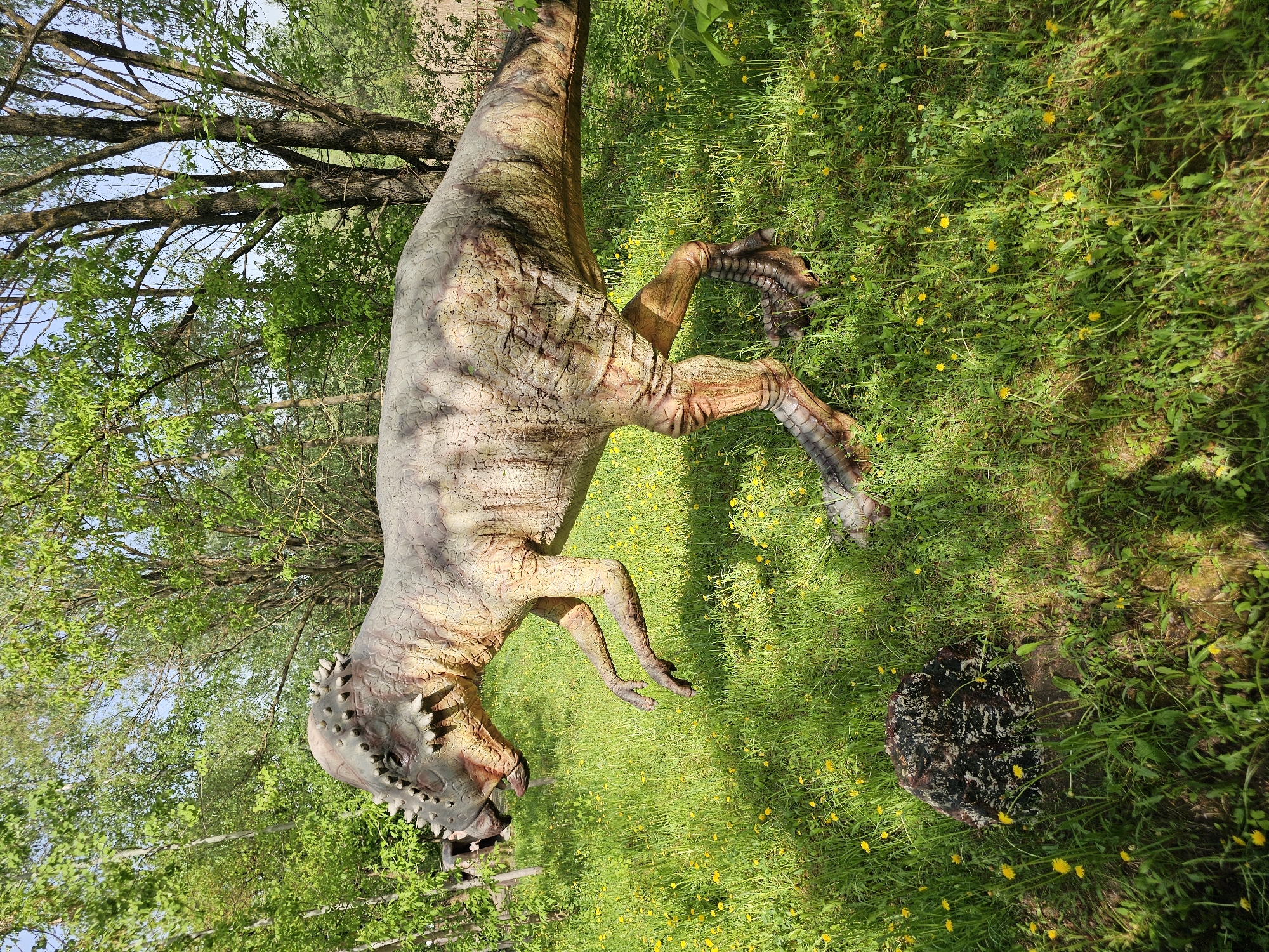 A dinosaur statue stands in a grassy area dotted with yellow flowers. The statue is brown and tan, with a textured skin pattern. Trees surround the statue. This location is in Khimki, Russia.