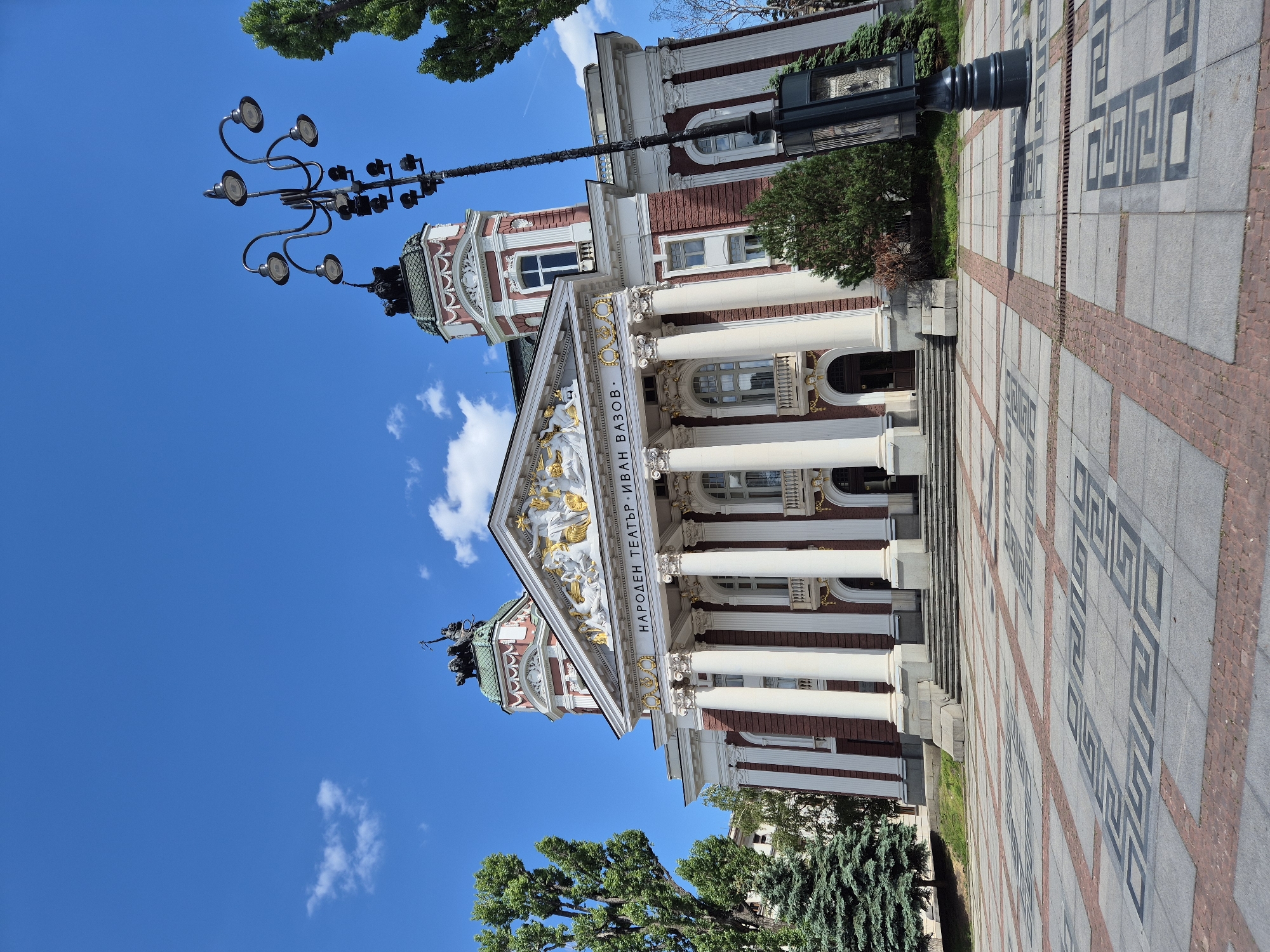 Here's a description of the image:
The image features the Ivan Vazov National Theatre in Sofia, Bulgaria. The building is a grand structure with white columns, a red and white facade, and a triangular pediment adorned with golden sculptures. The name of the theatre is inscribed in Cyrillic letters above the entrance. A bronze sculpture group is positioned above the roof, likely depicting a winged figure with accompanying figures. The sky is bright blue with scattered white clouds, and the building is surrounded by trees. A lamppost with multiple ornate lights is visible near the building. The foreground showcases the pavement with a geometric pattern of grey and red tiles. The scene is captured in daylight under clear weather conditions.