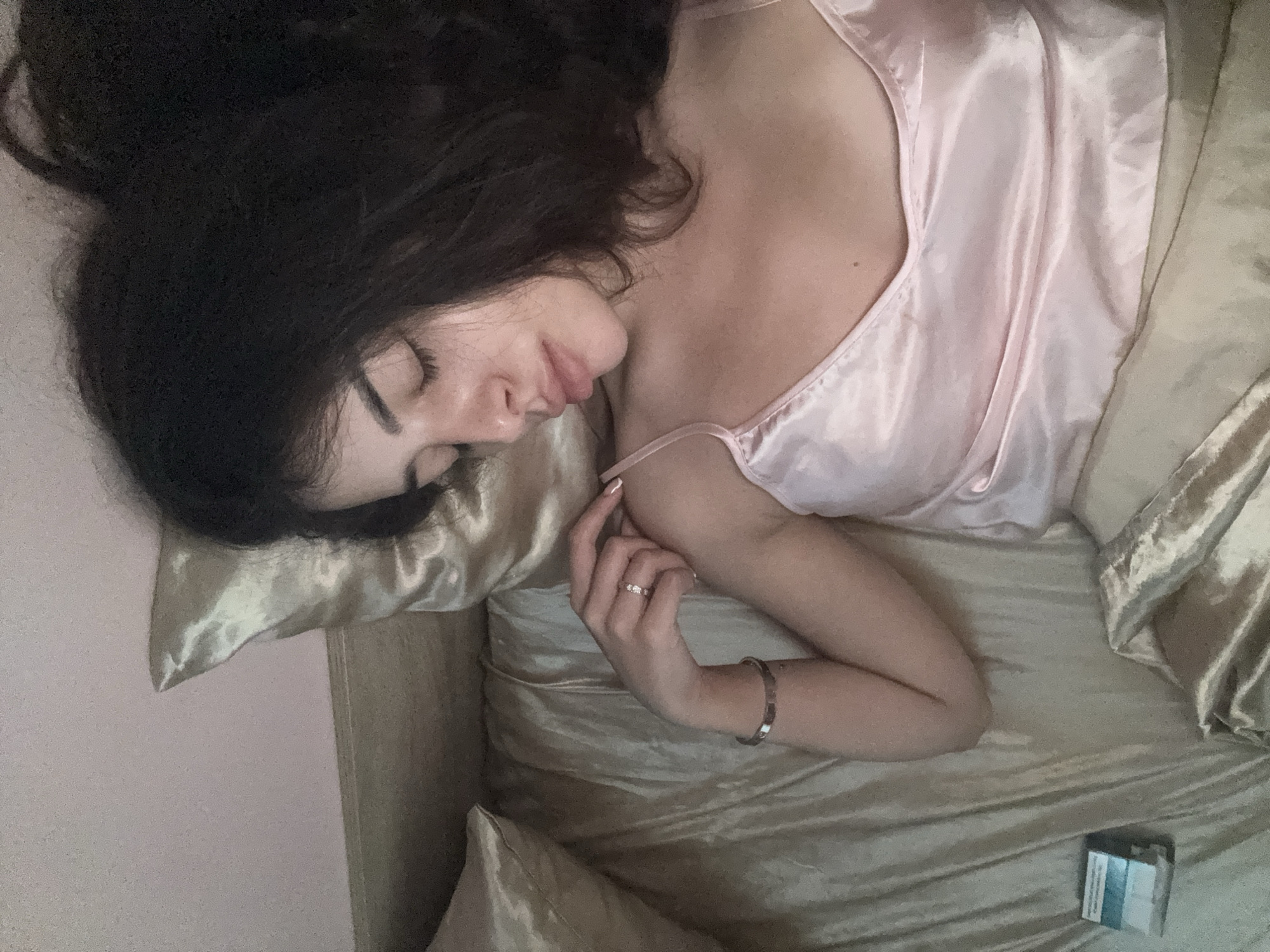 The image shows a young woman with dark hair lying on her side on a bed with satin sheets. She is wearing a pink satin camisole. Her eyes are closed, and she appears to be sleeping. Her right hand is resting on her chest, and she is wearing a ring on her left hand and a bracelet on her left wrist. To the left of the woman, on the bed, there is a small, rectangular blue and white object, which appears to be a pack of cigarettes. The background consists of a pink wall and a pillow. The lighting in the room is dim, suggesting it might be nighttime or early morning. The image does not provide any information about the location in Plovdiv, Bulgaria.
