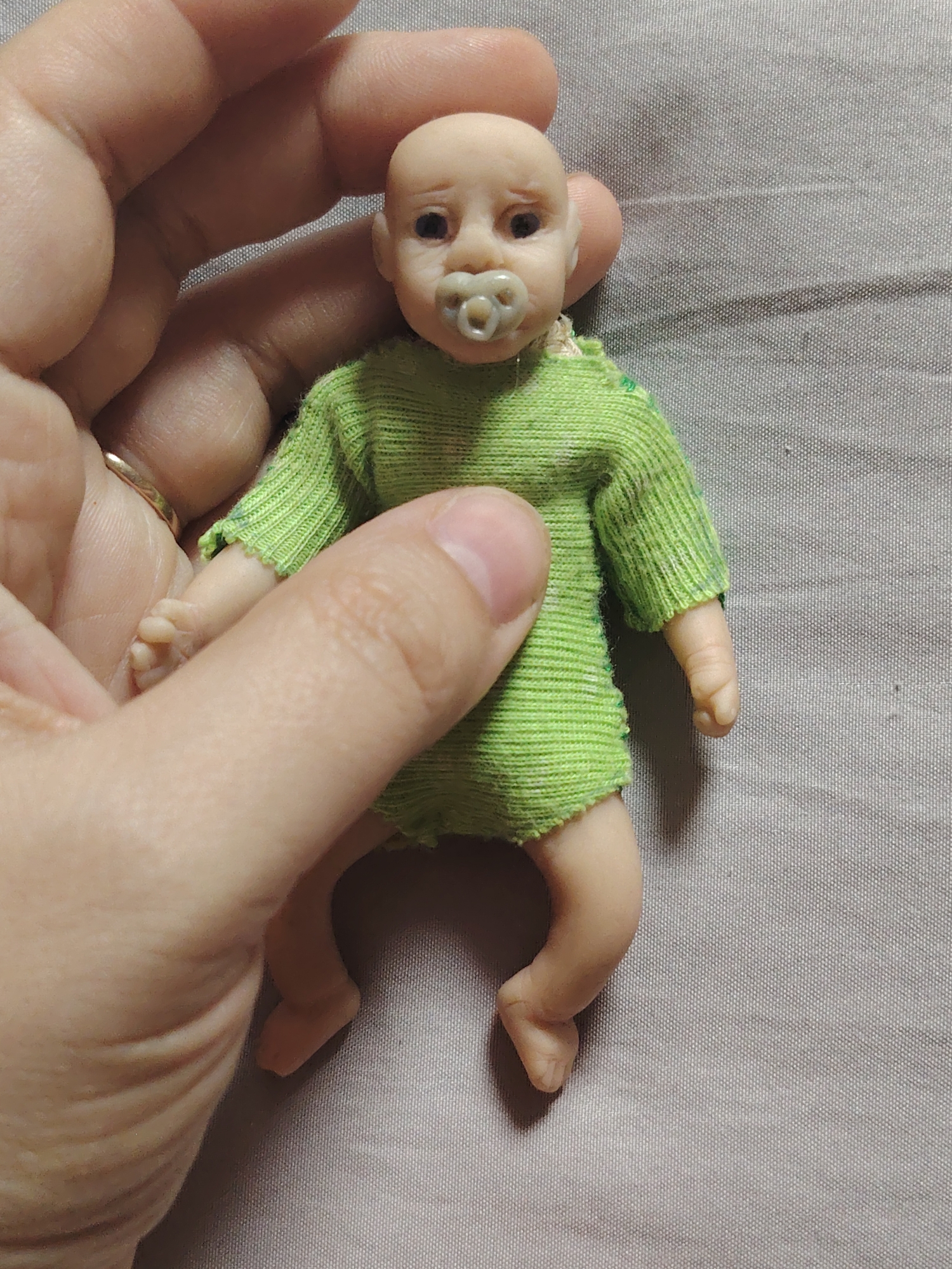 The image shows a close-up of a person's hands holding a miniature doll that resembles a baby. The doll is wearing a green, ribbed garment and has a pacifier in its mouth. The background is a textured grey fabric. The lighting is bright and appears to be artificial, suggesting an indoor setting. There is no visible text in the image. The hands are positioned around the doll, with fingers gently holding its body and feet. A ring is visible on one of the fingers. The doll's features are detailed, with a bald head, closed eyes, and small limbs. The overall impression is that of a close-up shot focusing on the doll and the interaction with the hands.