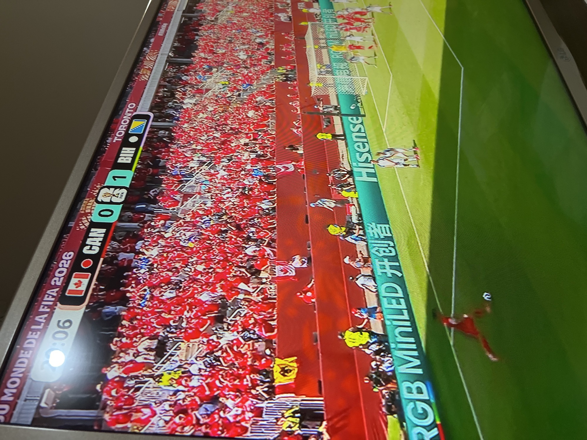 An outdoor electronic scoreboard at a soccer stadium displays game information. The scoreboard shows "JU MONDE DE LA FIFA 2026" at the top. Below this, the time reads "29:06" and the score is "0-1". The teams are represented by flags and abbreviations: a Canadian flag and "CAN" are on one side, and a Bosnian flag and "BIH" are on the other. The word "TORONTO" is also visible. The stadium seating is filled with a large crowd of spectators, most of whom are wearing red. The field is green grass with white lines. A soccer game is in progress, with players on the field near a goal. Several security personnel are also present. A banner with the words "RGB MiniLED 开创者" and "Hisense" is visible along the edge of the field. The image is taken from a slightly elevated angle, looking down at the field and scoreboard.