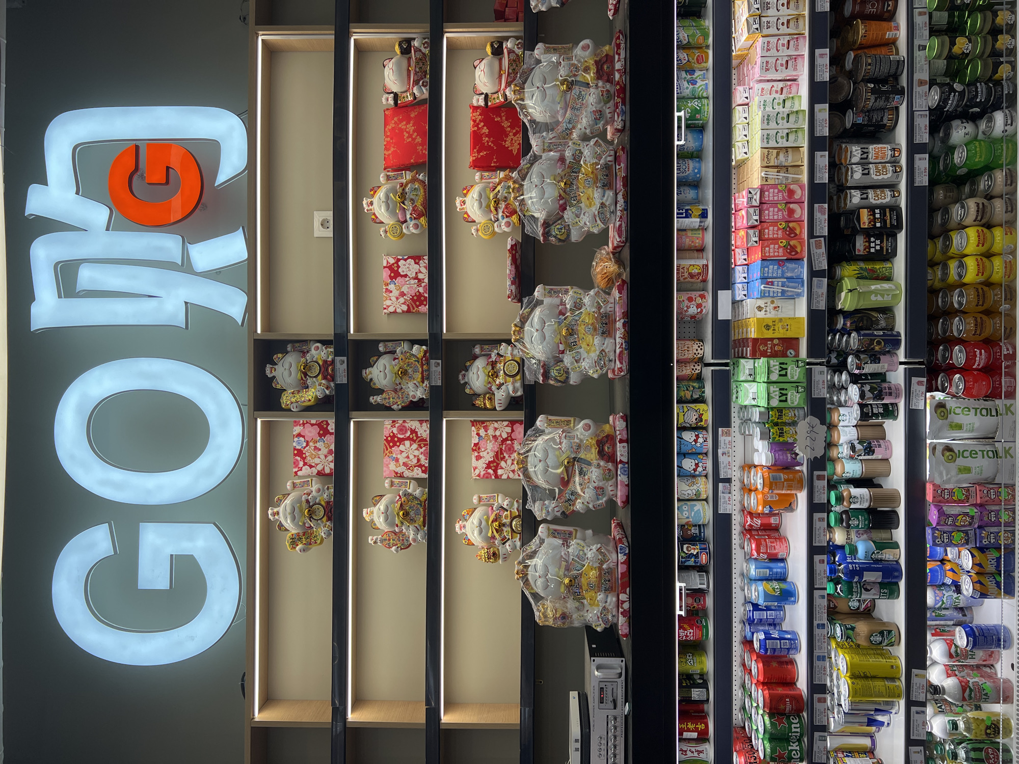 The image shows the interior of a grocery store, likely an Asian market, in Barcelona, Spain. The sign above the shelves reads "GO" in large, illuminated white letters, with an orange "G" overlapping the second character of the Chinese word "购" (gòu), which means "to buy." The shelves are well-lit and display various products.
The top shelves feature several decorative "maneki-neko" (beckoning cat) figurines, some small and intricate, others larger and wrapped in plastic. These are interspersed with decorative boxes and some food items.
Below the figurines, the shelves are filled with an assortment of beverages. The lower shelves are packed with rows of canned drinks, including energy drinks, sodas, and teas, with brands visible such as Starbucks, Hello Kitty, and various Asian beverage companies. There are also cartons of juice and milk-based drinks. The overall impression is that of a retail environment stocked with a wide variety of Asian food and beverage products.