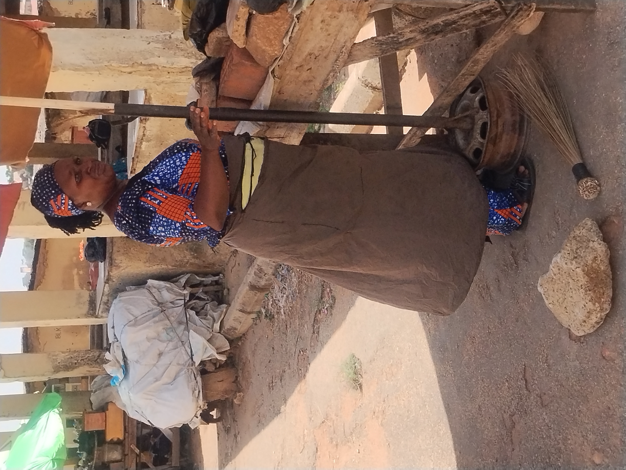 A woman stands outdoors in New Stadium, Nigeria, holding a horizontal metal bar with both hands, her palms facing each other. She wears a blue and orange patterned top, a long brown skirt with a yellow stripe near the waist, and a blue and orange patterned headscarf. Her feet are clad in blue patterned sandals. She stands next to a metal wheel and a broom. Behind her, there is a wooden structure supporting rocks and bricks. To the left of the woman, there is a large white bag, an umbrella, and assorted crates. The ground appears to be a mix of dirt and concrete.