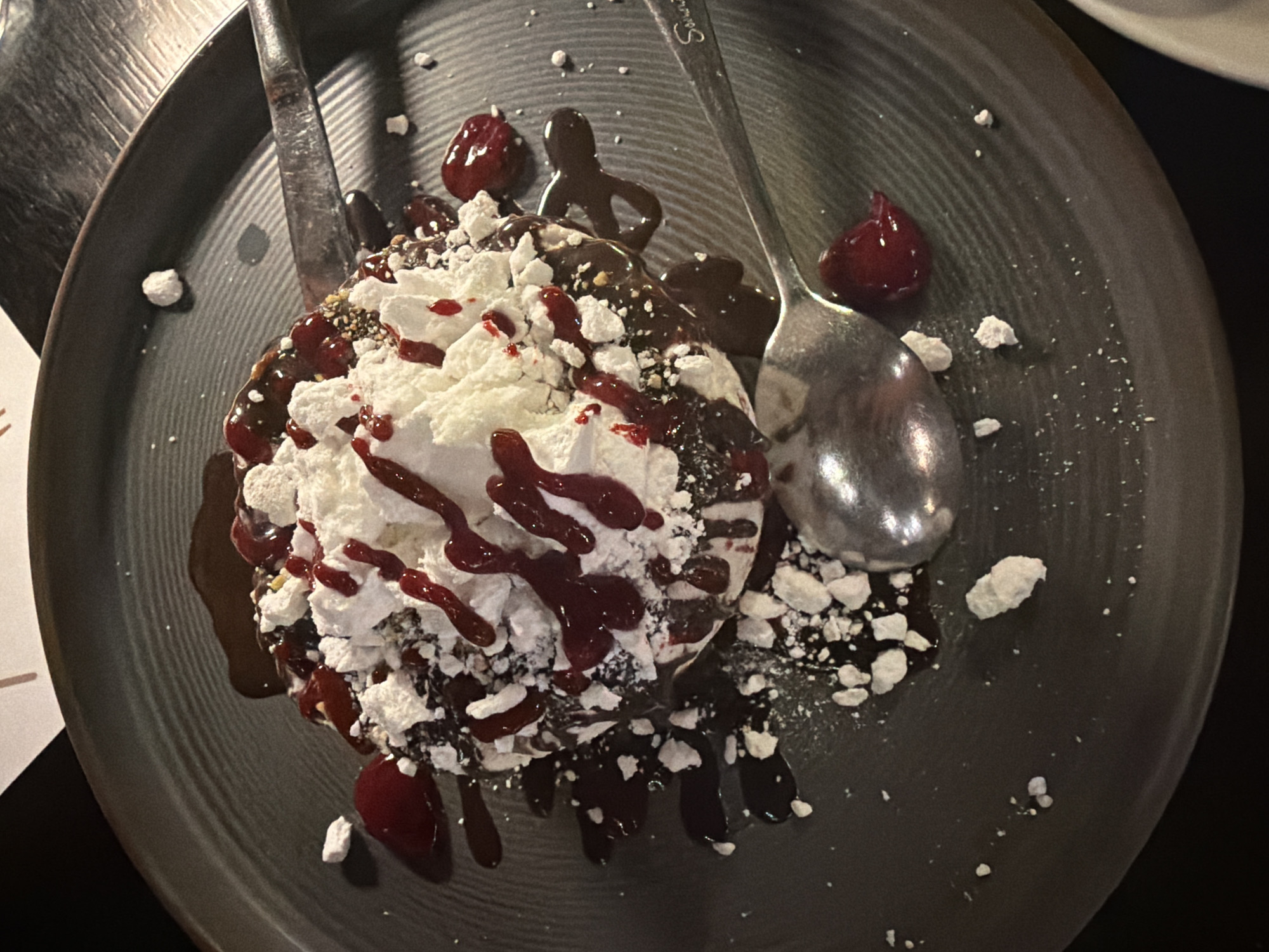 An overhead shot presents a decadent dessert on a dark grey, ridged plate. The dessert features a rich, dark chocolate base, possibly a brownie or cake, generously covered in a glossy dark chocolate sauce. A substantial amount of fluffy white topping, appearing to be whipped cream or crumbled meringue, sits atop the chocolate. This white topping is further adorned with scattered white meringue pieces and fine brownish sprinkles. A vibrant dark red berry sauce or coulis is drizzled extensively over the white topping and around the edges of the dessert. Several small dollops of berry compote or whole berries are also visible on the plate.
A silver spoon, with the faint word "Souvenir" visible on its handle, rests on the right side of the plate, partially submerged in the dessert. A silver fork is visible on the left, slightly out of frame. The plate itself has subtle concentric ridges, adding to the visual texture. The dim lighting suggests an evening setting, possibly in a restaurant in Makri, Greece.