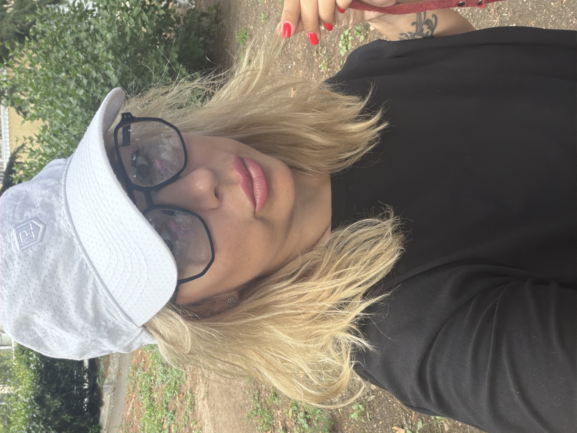A woman with blonde hair, wearing a white baseball cap, black t-shirt, and sunglasses, is pictured outdoors. Her lips are painted pink. She is holding a red leash in her right hand, and her fingernails are painted red. A tattoo is visible on her left arm. The background consists of green foliage and a gravel path.