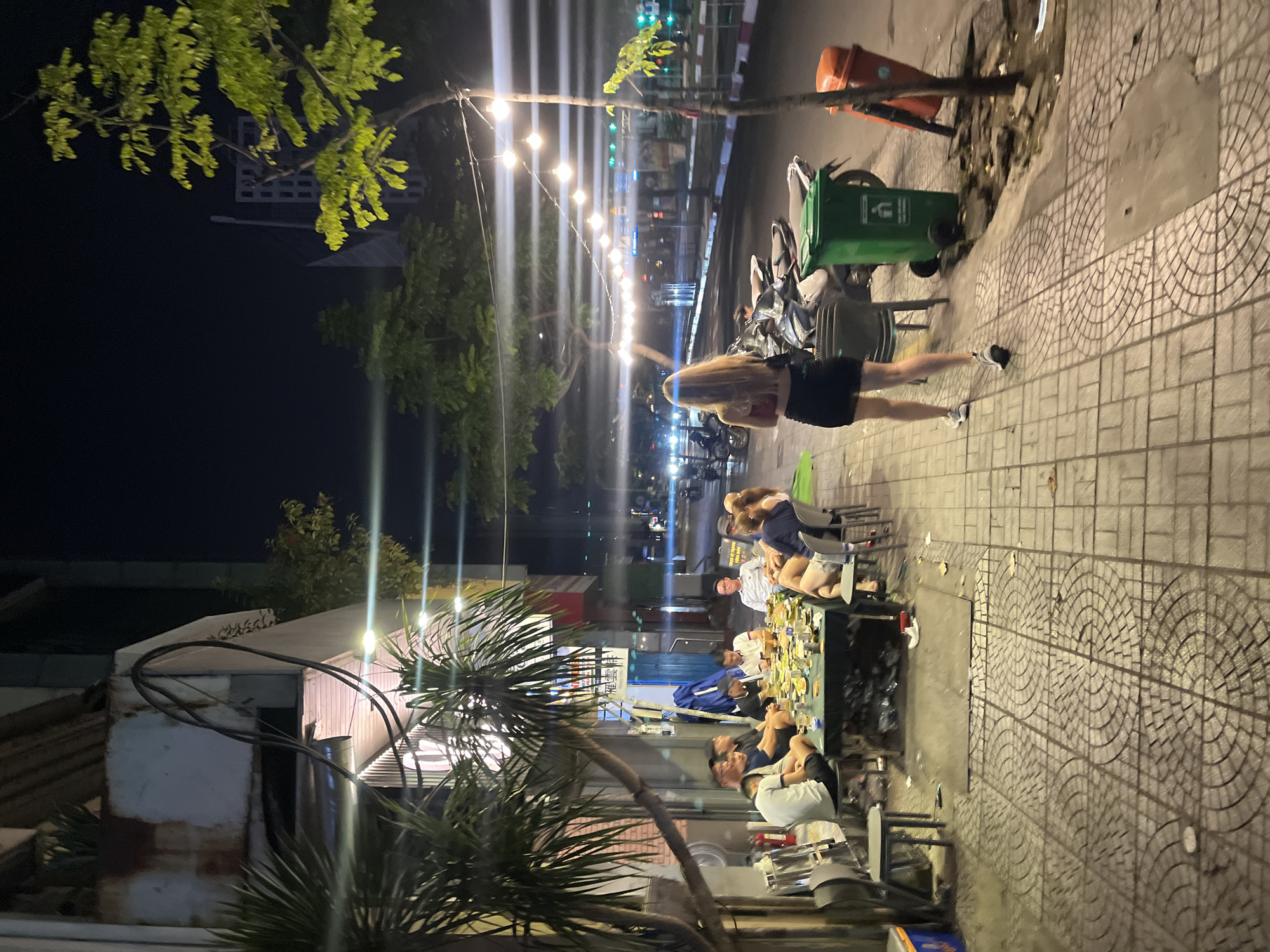 Captured at night in the city of Thủ Đức, Vietnam, the image presents a street scene. A group of people occupies an outdoor dining area, sitting around a table filled with food and drinks. A woman in a short skirt and blonde hair walks past on the sidewalk. Overhead, string lights illuminate the street. Street furniture includes trash bins, while parked motorbikes line the curb. A tree is in the foreground. Buildings are visible in the background.