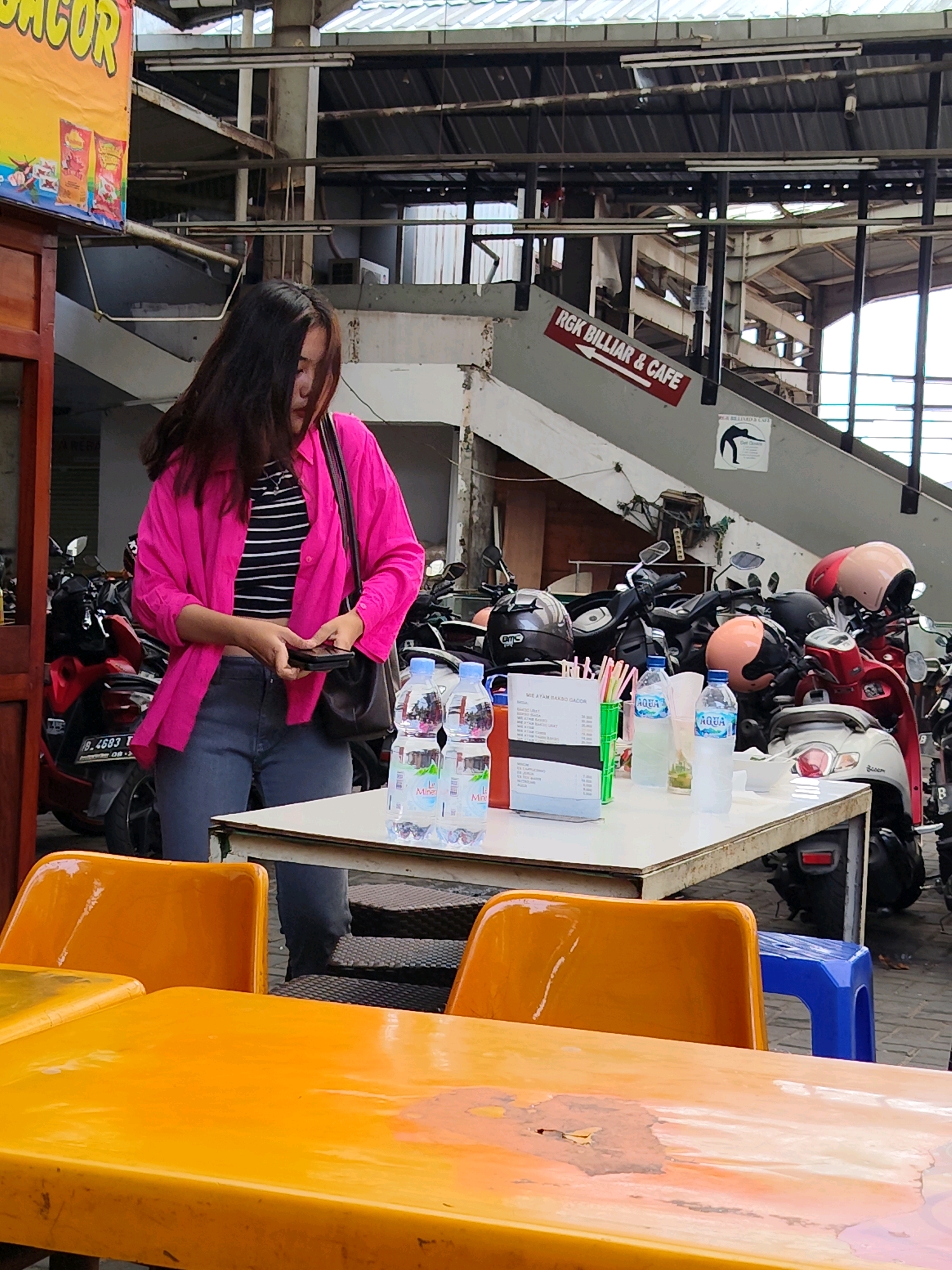 A young woman with dark, shoulder-length hair stands near a white table laden with drinks and a menu in what appears to be an outdoor seating area. She is wearing a bright pink oversized button-down shirt over a black and white striped crop top, dark gray jeans, and carries a dark purse over her left shoulder. She is looking down at her hands, which are holding a black smartphone.
The table is set with two bottles of Aqua brand mineral water, several drinking straws, and a menu displaying prices in Indonesian Rupiah. A stack of white bowls sits behind the drinks. In the background, numerous motorcycles are parked closely together, some adorned with colorful helmets. To the left of the woman, a red scooter is visible.
Above the motorcycles, a metal roof structure is supported by beams, suggesting an open-air market or a parking area with a canopy. A sign with an arrow pointing left reads "RGK BILLIARD & CAFE" in red lettering against a white background. Another poster featuring a silhouette of a person playing billiards is affixed to a concrete support. The overall environment suggests a bustling urban location, possibly a food court or a leisure area connected to a billiard hall. The presence of the Aqua brand water and the Indonesian currency on the menu strongly indicate the location is in Indonesia. Given the typical urban density and infrastructure seen, Central Jakarta is a plausible setting.