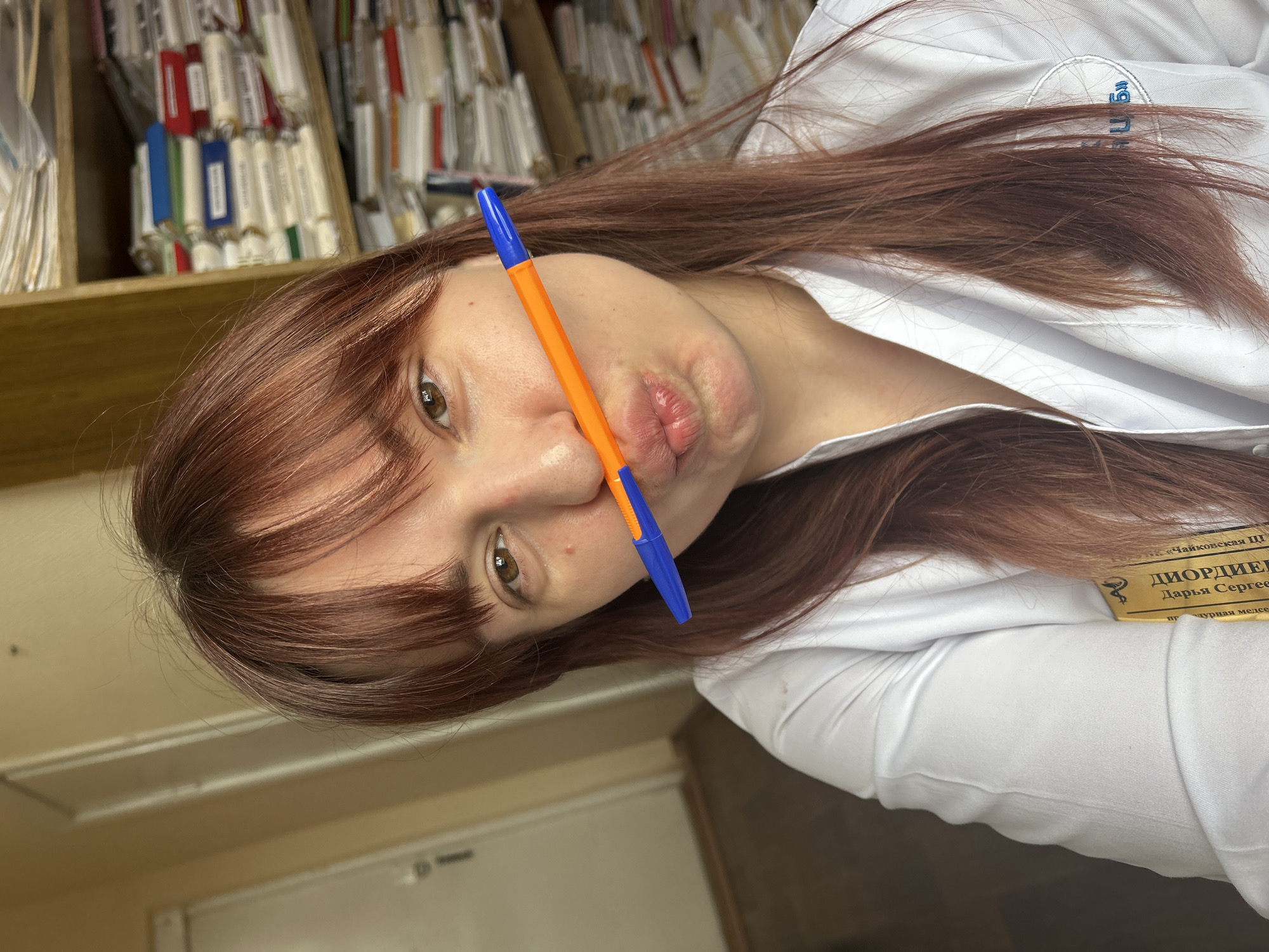 The image shows a woman with auburn hair, wearing a white medical coat with an identification badge, holding an orange and blue pen between her nose and lips, and posing for a selfie. The badge has text in Russian, including "Чайковскои ЦГБ" and the name "Диордиева Дарья Сергеевна", indicating she may be a medical worker at a hospital in Chaykovsky, Russia. Behind her is a shelf stacked with medical records or files. A door with a dark handle is also visible. The scene gives the impression of a break or moment of levity within a clinical environment.