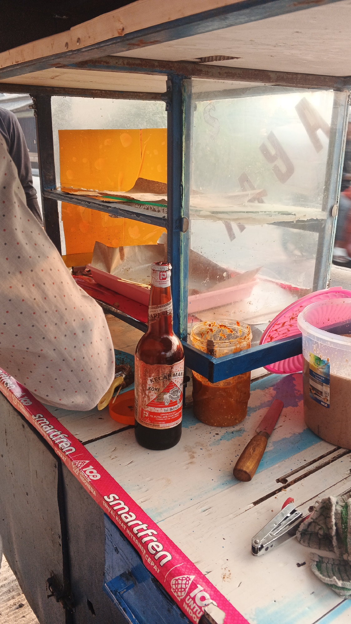 The image depicts a close-up view of a food cart or stall. In the foreground, a wooden surface is covered with various items, including a bottle of "KECAP MANIS KOKI" sauce, a small jar filled with a red, textured condiment, and a knife with a wooden handle. A pink promotional banner for "smartfren" runs along the edge of the cart. To the left, a portion of a person's torso is visible, draped in a white cloth with a pattern of small red dots. Behind these items, a glass-enclosed section of the cart holds more containers and what appears to be orange colored material on a shelf. The overall scene suggests a street food vendor's setup. The background is blurred, showing hints of an outdoor environment, possibly a street or market.