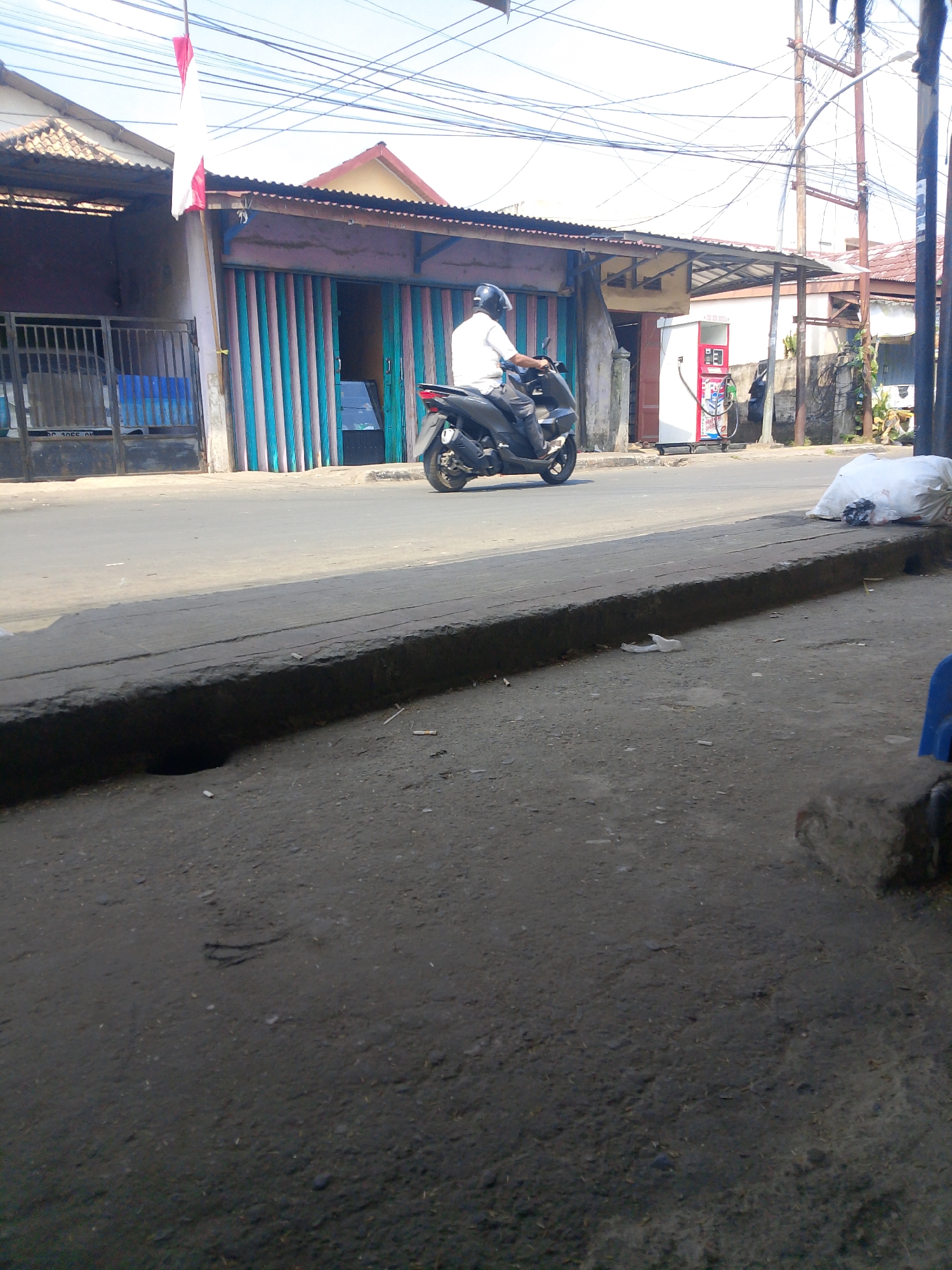 The image depicts a street scene in Palembang, Indonesia. In the foreground, a concrete curb runs along the side of a paved road. The road appears to be an urban thoroughfare with some scattered debris. On the opposite side of the road, there are several buildings with corrugated metal roofs and shutters. One building has a red and white Indonesian flag hanging from a pole. A person is riding a scooter on the road, wearing a helmet. To the right of the buildings, there is a white vending machine with a red accent and a hose attachment. A large white bag of what appears to be trash is placed on the sidewalk near the vending machine. The sky is visible in the background, with telephone wires crisscrossing above the street. The overall impression is a snapshot of everyday life in an Indonesian city.
