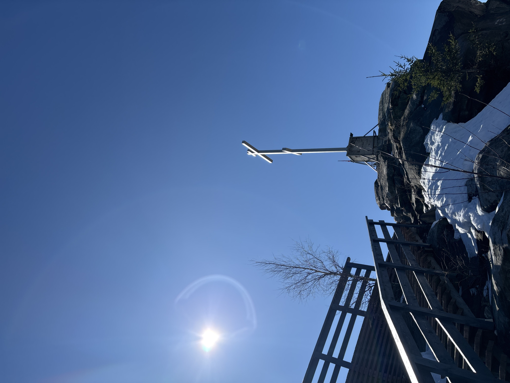 The image depicts a low-angle view of a rocky terrain on a sunny day in what appears to be the city of Gubakha, Russia. A white cross stands atop a rock face. A wooden structure, possibly a staircase or walkway, ascends the rock face. Snow is visible on the rocks, indicating a cold climate. The bright sun is in the lower part of the frame, creating a lens flare effect. The sky is clear blue.