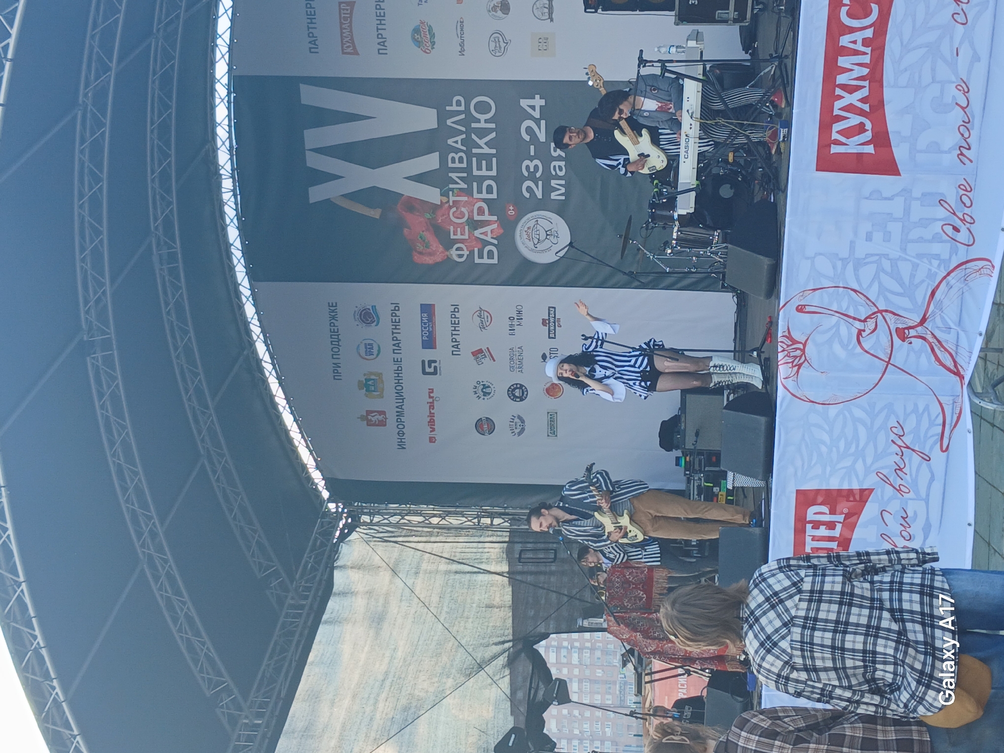A live band is performing on an outdoor stage at the XV Festival of Barbeque in Yekaterinburg, Russia, held May 23-24. The stage features large banners with event branding, including the "Kuhmaster" logo. The band consists of a female singer, a keyboard player, and two guitarists dressed in striped shirts. The crowd, including some in plaid shirts and jeans, is watching the performance. Supporting sponsor banners frame the top of the stage, while the scaffolding structure above is visible.