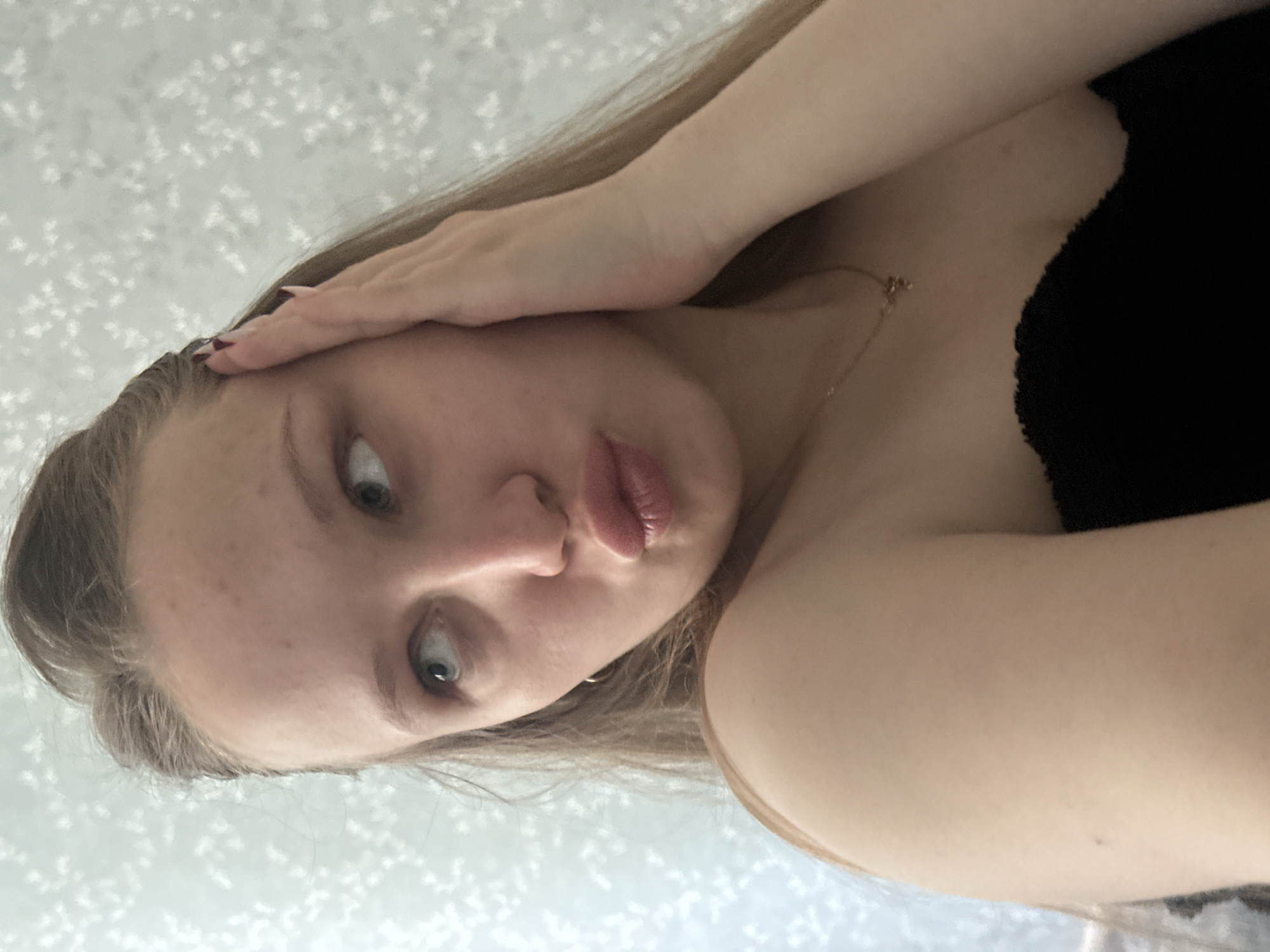 A fair-skinned woman with blonde hair and light eyes is pictured in a close-up selfie. She is holding one hand to her forehead. She is wearing a black top and a delicate gold necklace. The background consists of a patterned light-colored wall. The location in the city of Chaikovsky, Russia cannot be determined from the image.