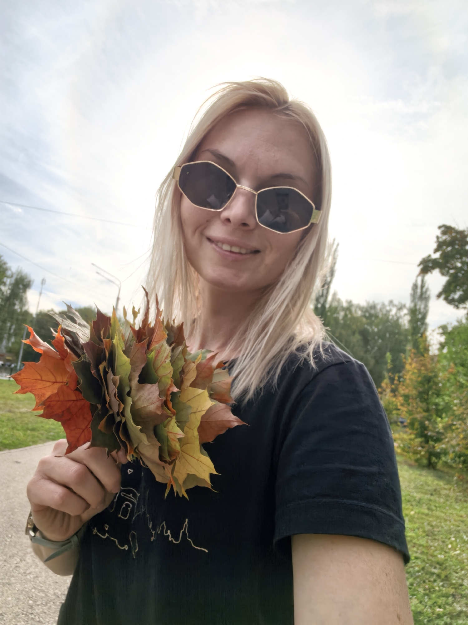 A young woman with blonde hair and sunglasses holds a bouquet of autumn leaves. She is wearing a black t-shirt and a watch on her left wrist. She is standing outdoors with trees and a cloudy sky in the background. The location is likely Sterlitamak, Russia, given the context.