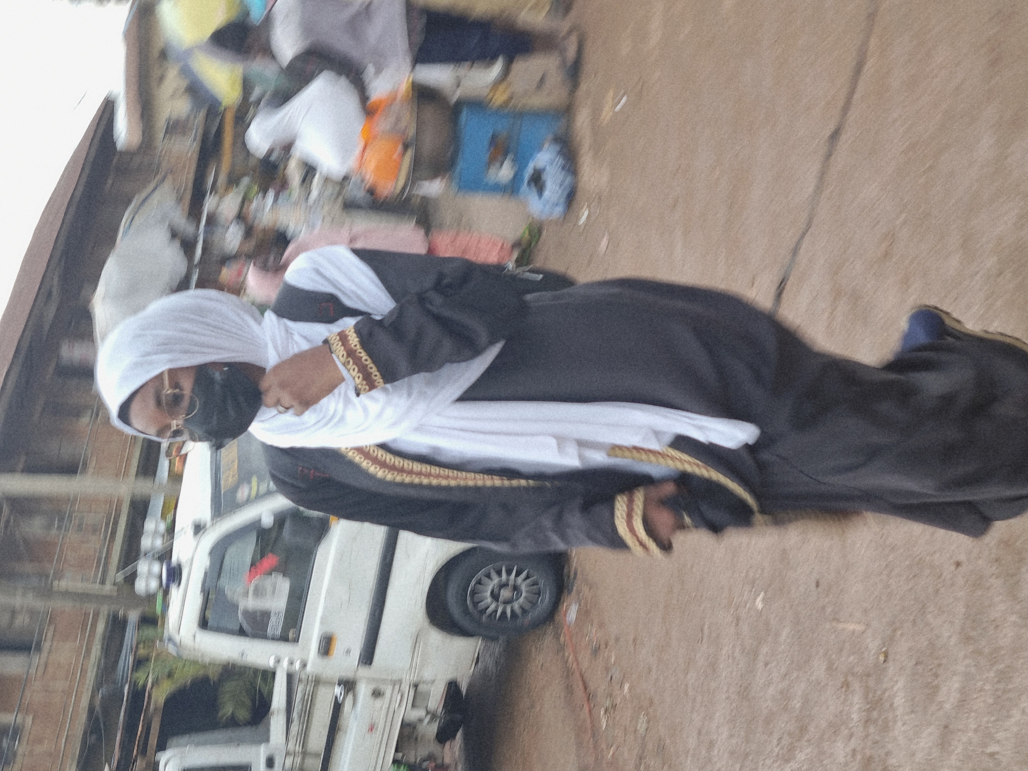 The image depicts a woman wearing a white hijab, black face mask, glasses, and a long black abaya with gold trim. She is walking on a dirt road, possibly in a market setting. A white truck with a covered bed is parked to her left. In the background, blurred figures of other people, market stalls, and buildings are visible, suggesting an urban environment. The lighting suggests an overcast day. The scene appears to be in Jos, Nigeria, a city known for its markets and diverse population. The woman's attire and the presence of a market suggest a cultural context typical of Jos. The overall image is slightly motion-blurred, indicating movement.