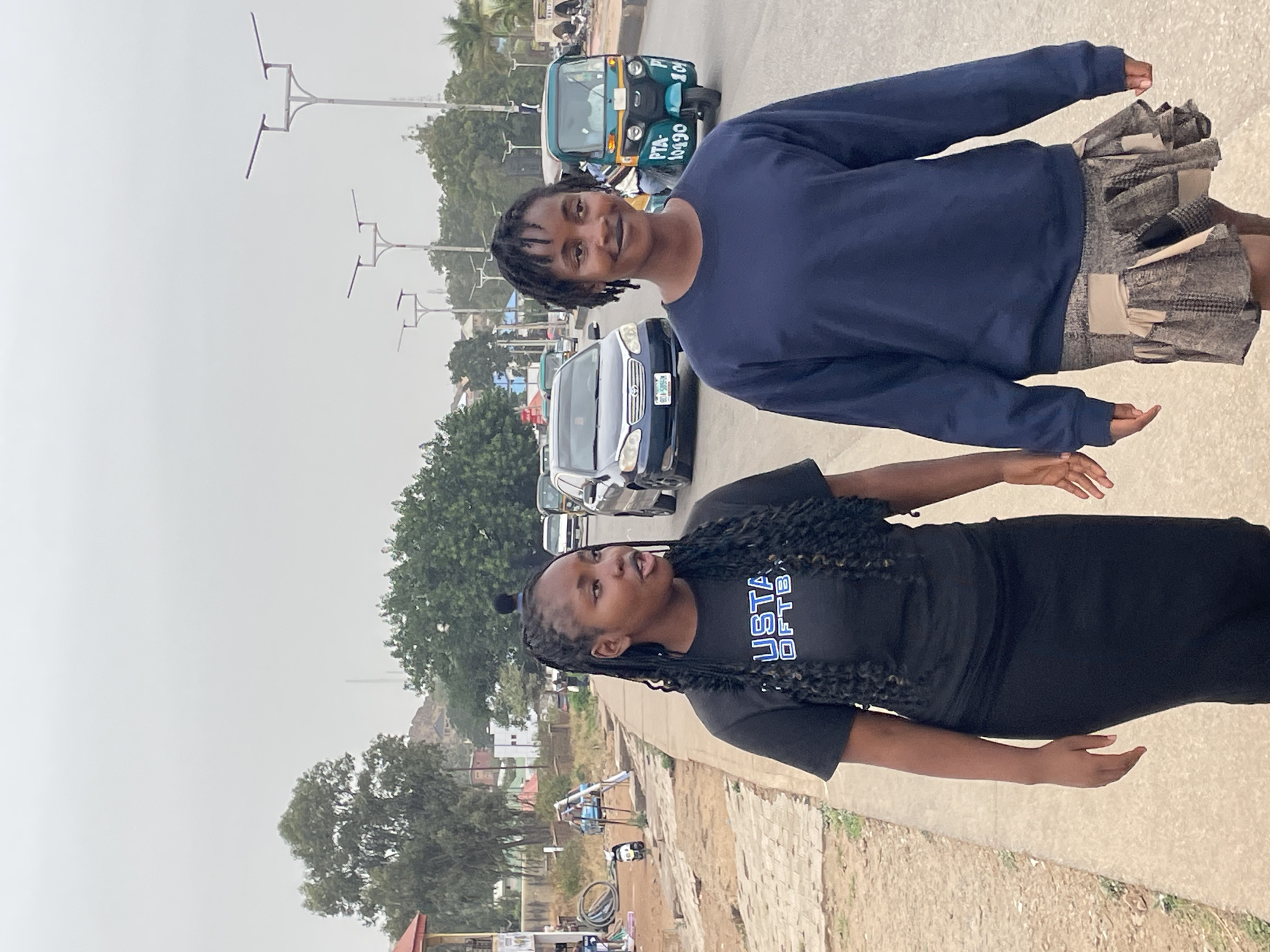 Two young women stand in front of traffic on an asphalt road. The woman in the foreground wears a black t-shirt with white and blue letters and a long black skirt. The second woman wears a navy blue long sleeve shirt and a multi-color, patchwork-style skirt. Behind them, light and dark colored cars and a blue three-wheeled vehicle are visible on the road. Green trees are in the background, and utility poles line the street. The scene is set on a cloudy day in Jos, Nigeria. A dirt area is visible in the foreground with a partially obscured bicycle, and a low wall separates the dirt area from the road.