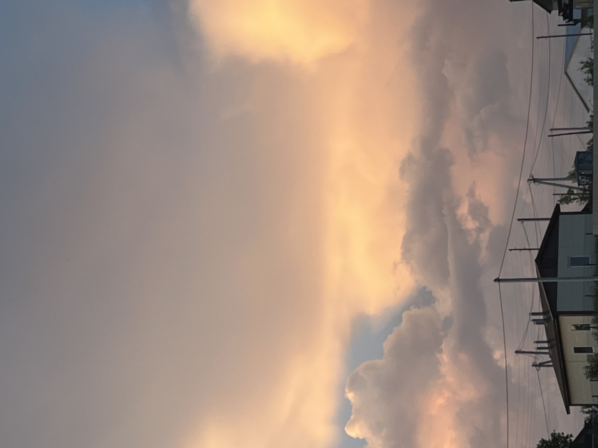 The image depicts a twilight sky over a rural settlement, likely Ludorvay, Russia. The sky is filled with dramatic, billowing clouds, tinged with soft orange and pink hues from the setting sun. Below the vibrant sky, a low-lying building with a light yellow and white facade is visible, featuring several windows. Power lines and utility poles traverse the scene horizontally, suggesting a functional, residential area. The overall atmosphere is serene and natural, illuminated by the gentle light of dusk.