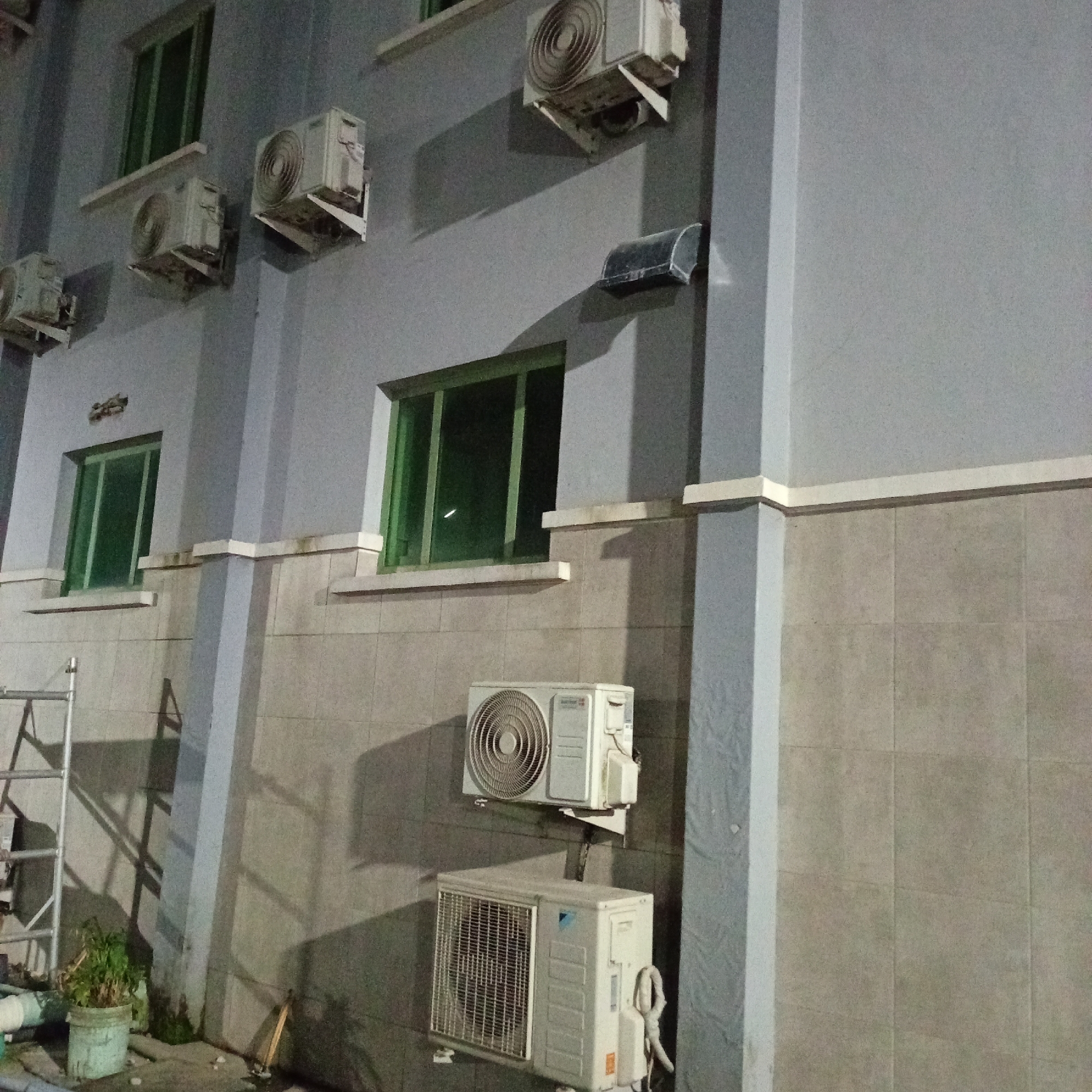 The image shows the exterior wall of a building with multiple air conditioning units mounted on it. The building appears to be in an urban setting, possibly in Ibadan, Nigeria, given the context. The wall is painted a light gray, and there are several windows with green tinted glass. Some of the air conditioning units are older models, while others appear to be more modern. There is a ladder leaning against the wall on the left side of the image, and a potted plant is visible at the base of the ladder. The overall scene suggests a commercial or residential building that has undergone several installations of air conditioning systems. The lighting indicates it is nighttime or early morning.