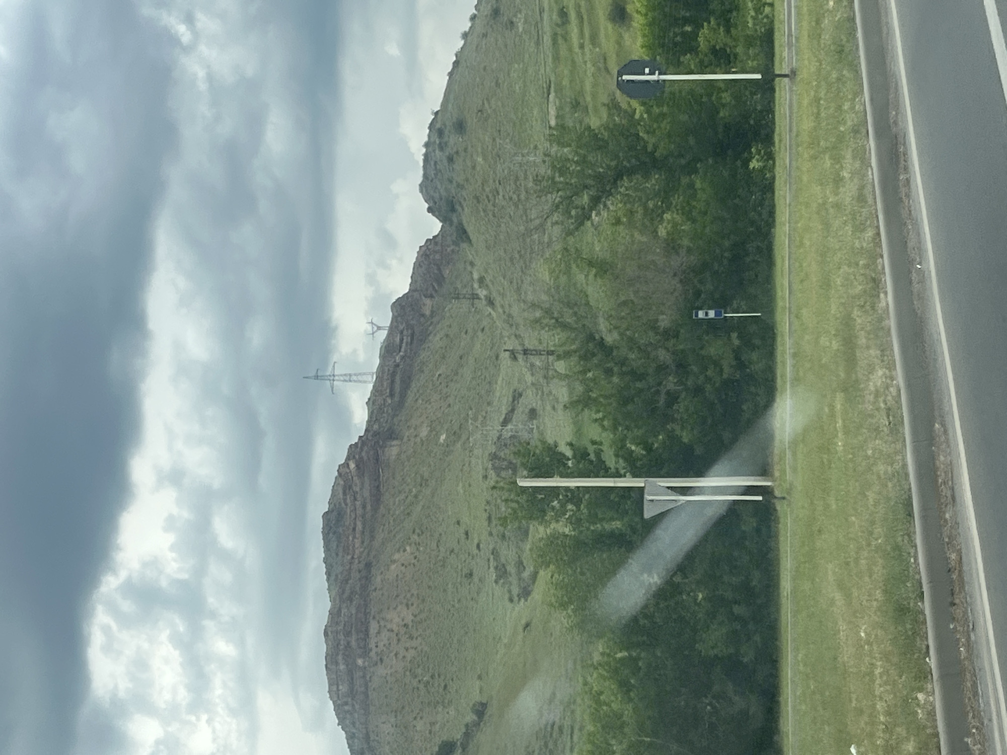 The image displays an outdoor landscape featuring a prominent mountain, a road, and an overcast sky, likely taken from a moving vehicle.
A large, rugged mountain or steep hill dominates the background. Its slopes are mostly covered in green vegetation, primarily sparse grass and shrubs, with rocky outcrops visible, especially along the ridge and lower sections. Several tall, lattice-style electricity transmission towers (pylons) with accompanying power lines are visible climbing and traversing the mountain's contours.
At the bottom of the frame, a paved road, likely an asphalt highway, stretches horizontally, featuring at least two lanes and a shoulder. A strip of green grass and dense green trees separate the road from the base of the mountain. Two roadside signs are visible near the trees. One is a black circular disc mounted on a white pole, partially obscured by foliage. The other, further along the road, is a smaller blue rectangular sign on a white pole, too blurry to discern specific details.
The sky, occupying the upper left portion of the image, is heavily overcast with varying shades of gray clouds, indicating cloudy weather and a subdued daytime light. A diagonal streak of light or glare is present across the bottom right, suggesting the photograph was taken through a window or windshield. The scene portrays a natural landscape integrated with infrastructure, located in Chiva, Armenia. No people or explicit activities are visible.