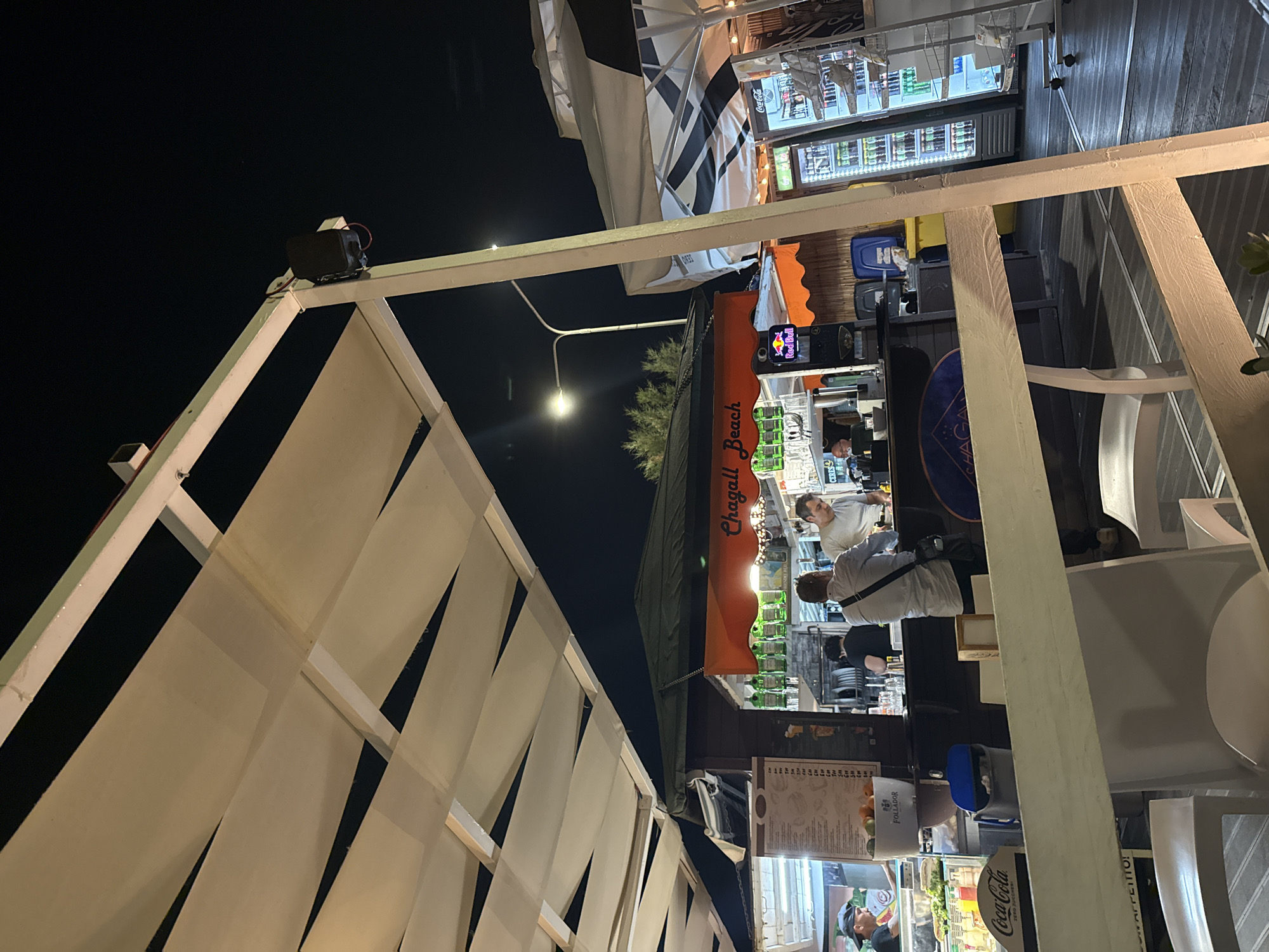 Captured at night in Capaci, Italy, the scene shows the exterior of a bar or beach club named "Chagall Beach". The bar is partially obscured by a white, pergola-like structure with fabric slats. A street light is visible beyond the bar. Inside the bar, several people are present, including a bartender. The bar is stocked with alcoholic beverages and features a Red Bull sign. Adjacent to the bar are refrigerated beverage displays. A Coca-Cola advertisement is also visible. The overall environment suggests a relaxed, outdoor venue.