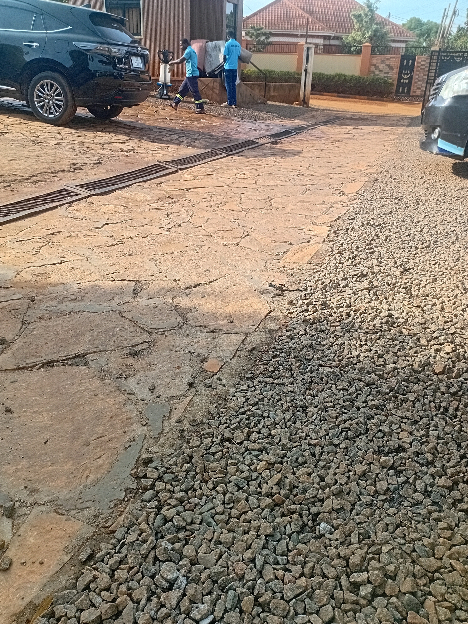 In Kira, Uganda, this image captures a scene at a car wash. Two men, dressed in blue shirts and dark pants, are seen working. One man is actively engaged, walking with a hose in his hand, suggesting he is washing a vehicle. The other man stands a short distance away. A black SUV is parked to the left of the frame, partially obscuring a metallic drain grate that runs across the ground. The ground itself is a mix of textured stone paving and loose gravel. In the background, a building with a corrugated iron facade is visible, and beyond that, residential housing with red-tiled roofs and fencing. A white car, possibly another vehicle being serviced, is partially visible on the right side of the image. The lighting suggests it is daytime, with shadows indicating the sun's position.