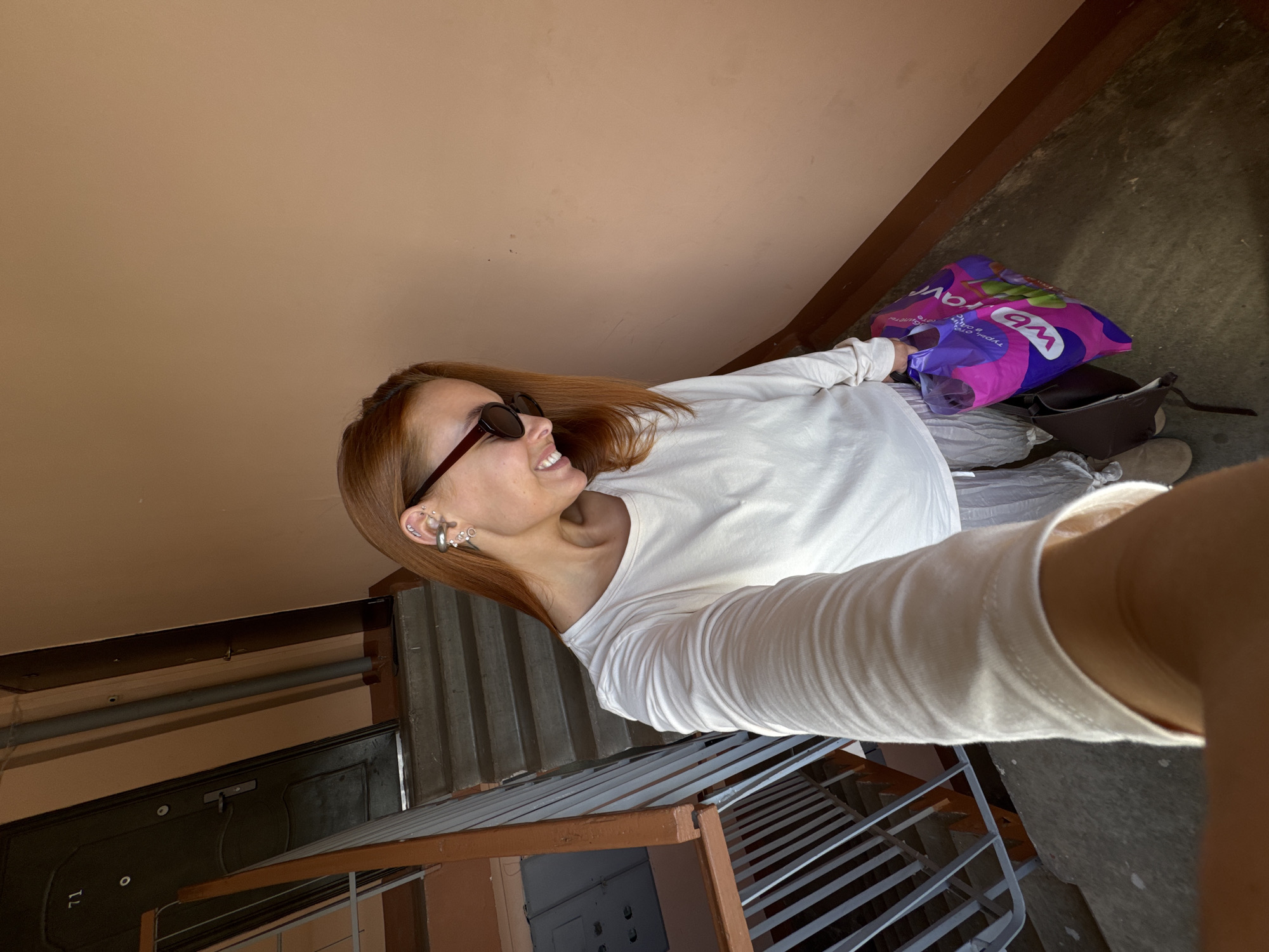 In Chaikovsky, Russia, a woman with auburn hair, wearing sunglasses and a white long-sleeved shirt, stands on an apartment building staircase, holding a purple shopping bag with the "Wildberries" logo. She is taking a selfie. The staircase has metal railings and concrete steps. The walls are a peachy-pink color. An apartment door with the number "71" is visible in the background.