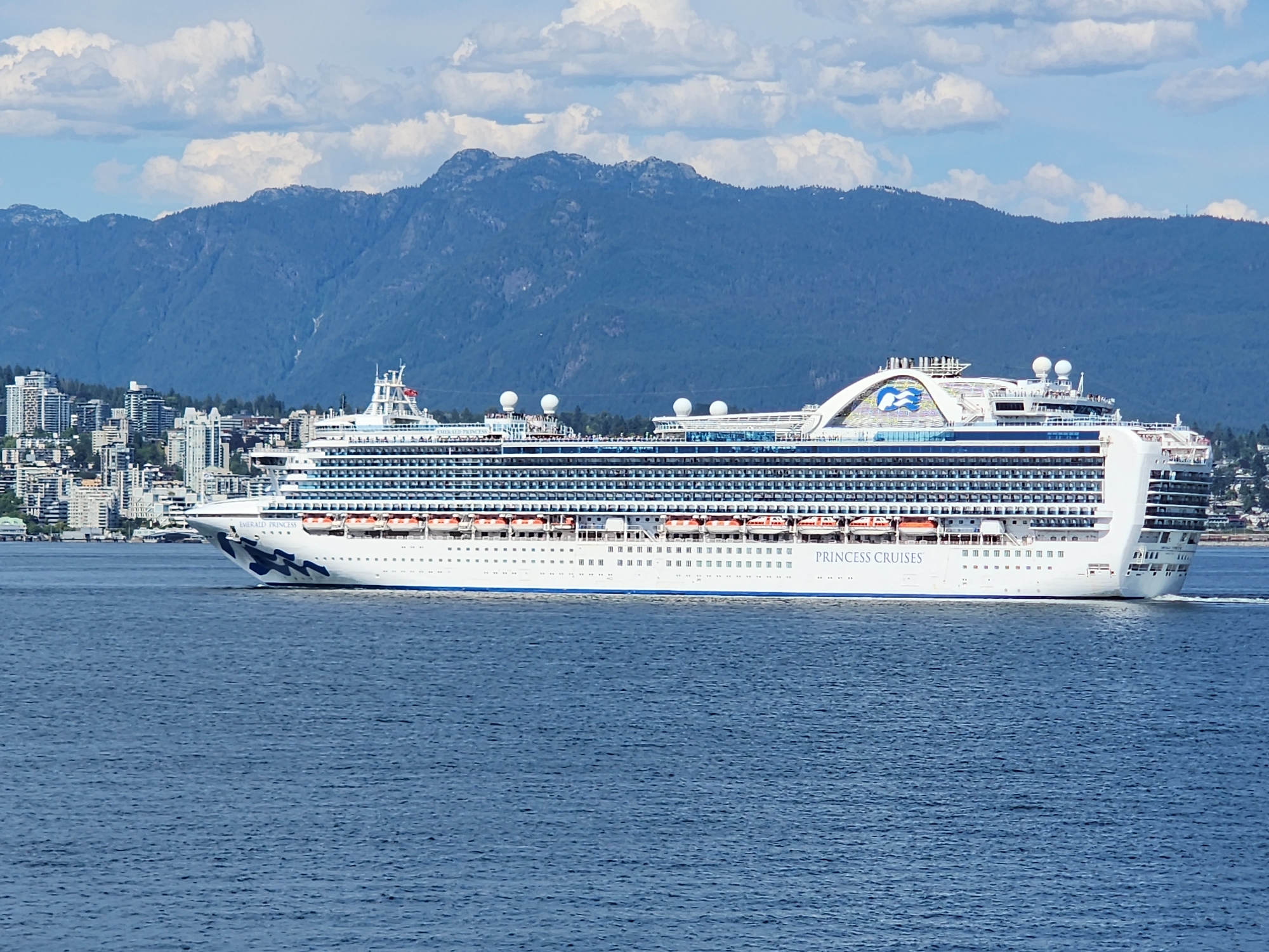 A large white cruise ship, the "EMERALD PRINCESS," is sailing on calm blue water. The ship has many decks with rows of windows and lifeboats visible. The Princess Cruises logo, a blue seahorse, is prominently displayed on the ship's superstructure. In the background, a city with numerous tall buildings is situated on the shore. Beyond the city, a range of blue-tinged mountains rises, with some white clouds scattered across the sky. The scene appears to be set during the daytime, under fair weather conditions, with bright sunlight illuminating the ship and the landscape. The water is a deep blue, reflecting the sky. The overall impression is one of a peaceful, scenic journey on a clear day. The text "PRINCESS CRUISES" is visible on the side of the ship.