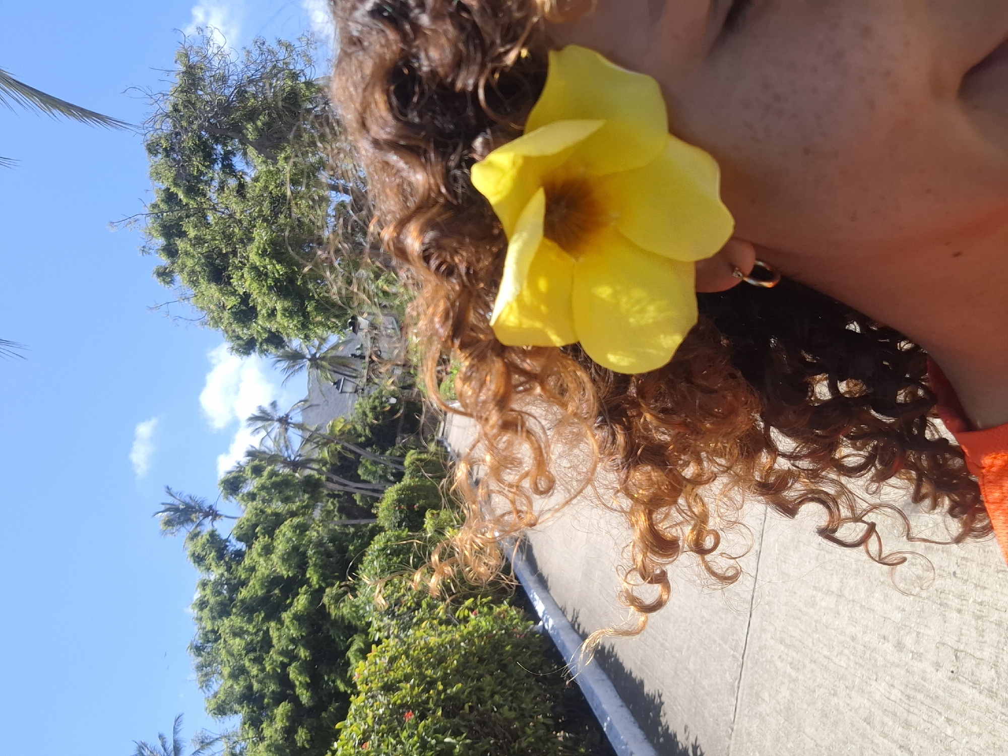 The image, oriented vertically, captures a close-up of a person's side profile, their head tilted towards the right. A prominent bright yellow flower is tucked behind their ear, adorned with a small, reflective hoop earring. The person has curly, reddish-brown hair, and their skin shows subtle freckles.
The background features a vivid blue sky, dotted with fluffy white clouds, occupying the upper and left portions of the frame. Below the sky and behind the person's head, dense green foliage of tropical trees, including visible palm fronds, suggests a warm, verdant environment. A light-colored paved surface, possibly a sidewalk or road, runs along the bottom and lower right edge of the image, marked by strong shadows indicating bright daylight.
The scene evokes a sunny day in a tropical location, consistent with Playa El Agua, Venezuela. No specific activity is discernible beyond the person being outdoors. There is no visible text.