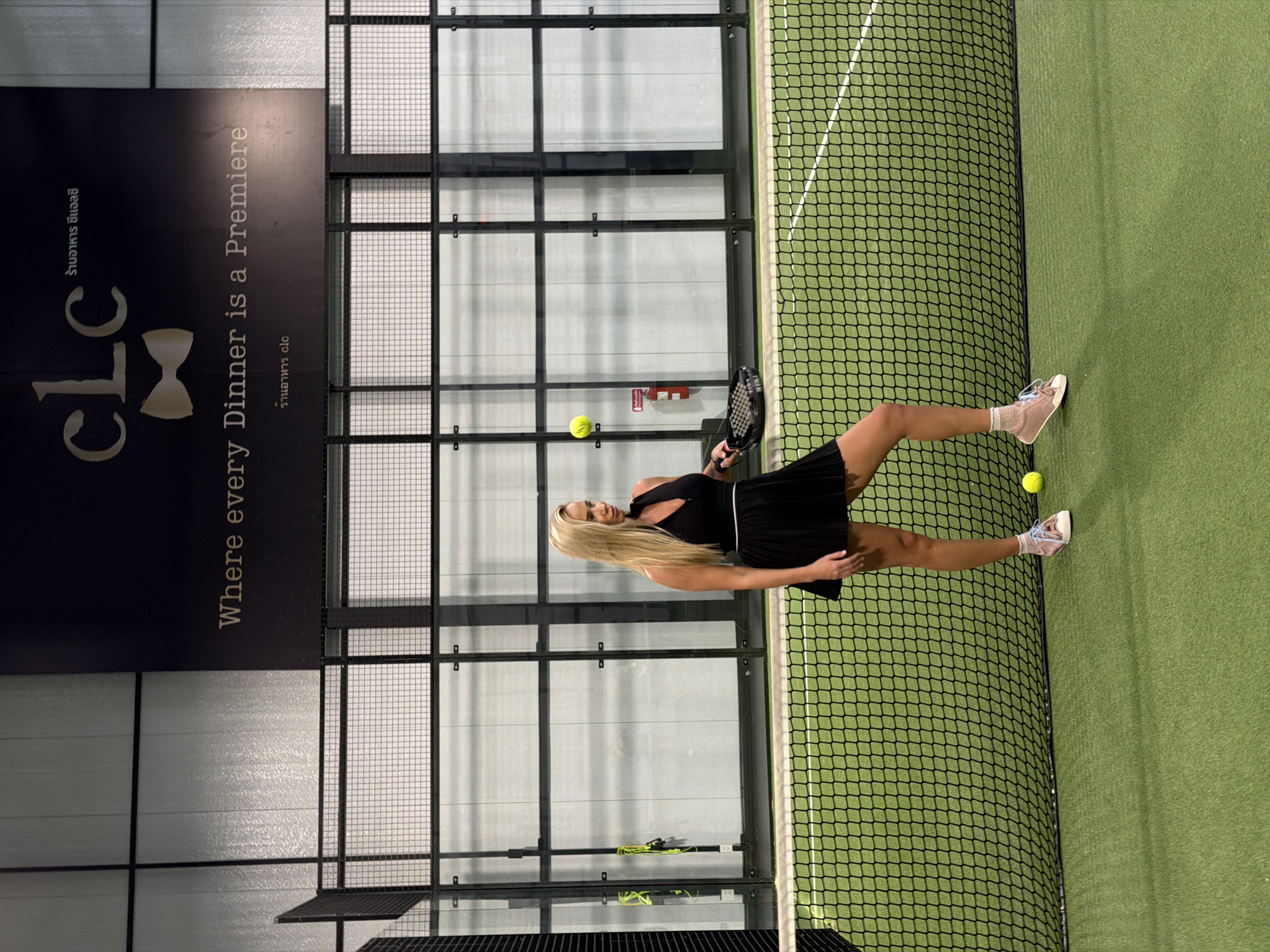 A woman with blonde hair is playing padel on an indoor court. She is wearing a black athletic outfit and pink sneakers. A yellow padel ball is in the air near her, and another is on the ground next to her. She is holding a black padel racket. The court has green turf and is enclosed by nets and glass walls. A large black banner with text is visible on the left side of the frame. The text on the banner includes "CLC" and "Where every Dinner is a Premiere." The overall lighting suggests it is daytime, possibly indoors with artificial lighting. The scene depicts a sports activity in a dedicated recreational facility.