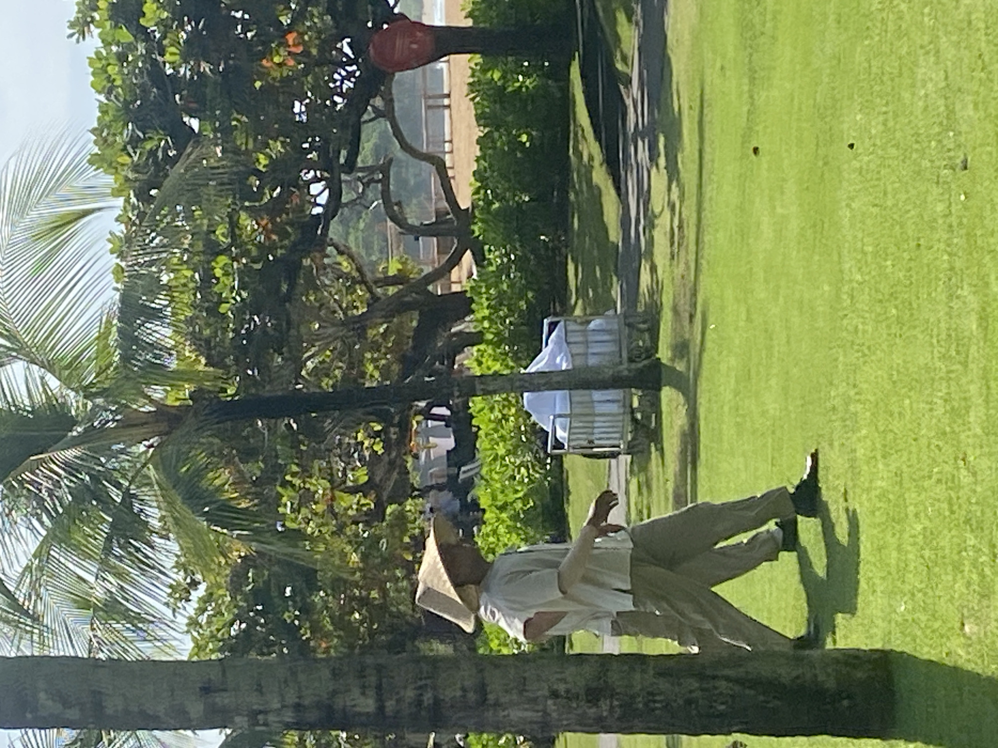The image shows a man on a grassy lawn, likely in Nusa Dua, Indonesia given the tropical vegetation and beach view in the background. The man wears a traditional Asian conical hat, a light-colored shirt, and khaki pants. He appears to be in motion, possibly walking or gesturing with his hands. Behind him, there are trees, including a palm tree, and a white cart is visible. In the distance, a beach and some structures are discernible. The scene is bathed in sunlight.