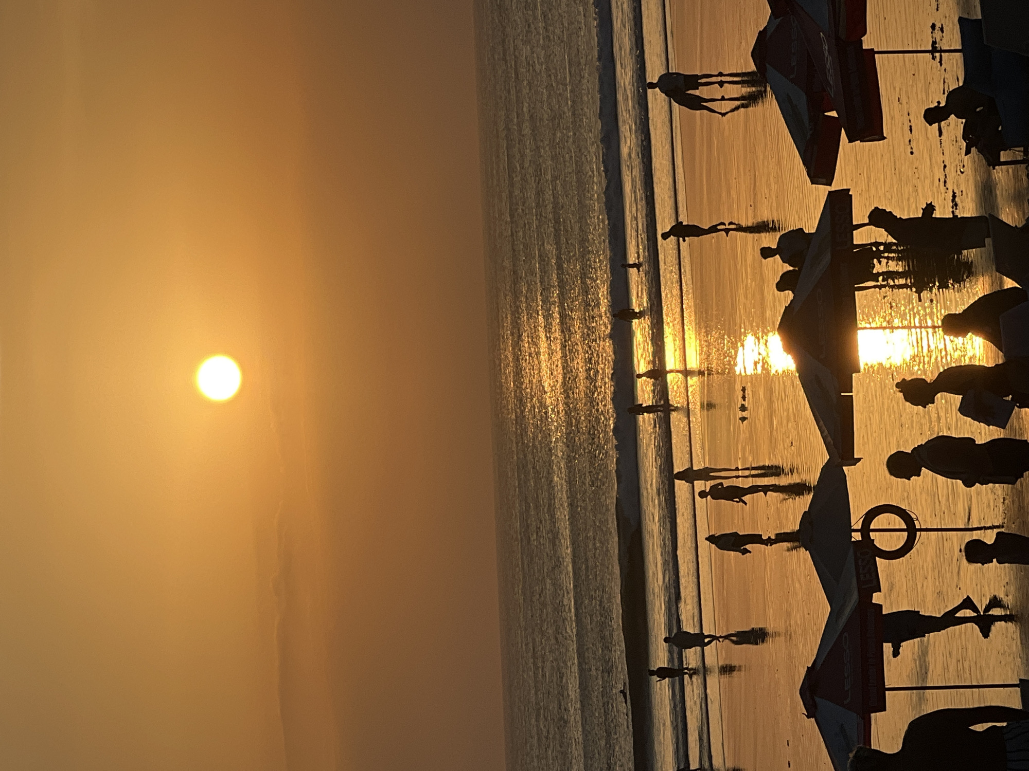 The image depicts a sunset on Kuta Beach in Bali, Indonesia. The sun is a bright orb in the upper center of the frame, casting a warm, golden light across the sky and reflecting on the wet sand and ocean. Silhouettes of people are scattered across the beach, some wading in the shallow water, others walking or standing. Umbrellas, likely from beach vendors, are visible in the foreground. The ocean displays gentle waves, and the reflection of the sun creates a shimmering path across the water's surface. The overall atmosphere is serene and idyllic, characteristic of a tropical beach at sunset.