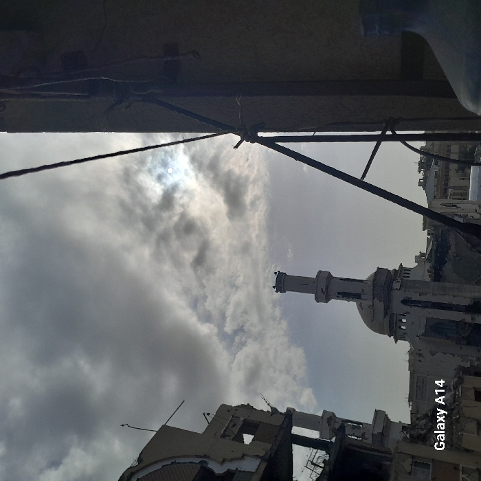 The image presents a dilapidated mosque with a prominent minaret against a cloudy sky. Buildings, possibly residential, show signs of significant damage and destruction, suggesting conflict or disaster. A metal structure with ropes is visible in the foreground, framing the scene. The overall impression is one of devastation and loss amidst a partially overcast sky.