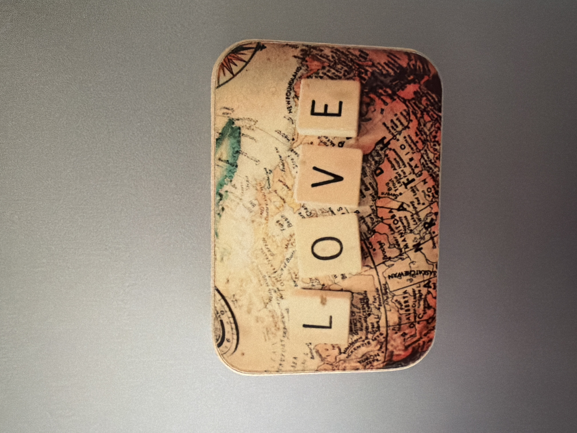 A close-up, eye-level shot captures a rectangular object with a vintage map design, likely a tin or decorative box. Four scrabble tiles spelling "LOVE" are placed on the map. The tiles are light-colored with black lettering. The map underneath is detailed with geographical features and place names, appearing to be of North America. The object is set against a plain, grey, textured background. There are no people or specific locations in Pazardzhik, Bulgaria visible in this image.