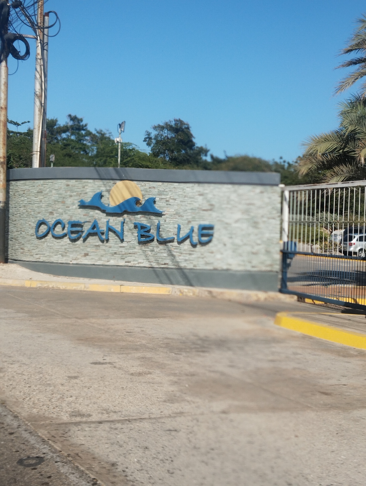 The image shows the entrance to "Ocean Blue" in Porlamar, Venezuela. The entrance consists of a low wall with the name "Ocean Blue" in blue lettering with a blue wave and a yellow sun above it. A gate stands slightly open to the right of the wall. A white vehicle can be seen through the gate. The road in front of the entrance is made of gray concrete. Yellow curbs delineate the road's edge. A clear blue sky is visible above. Trees and utility poles are present in the background.