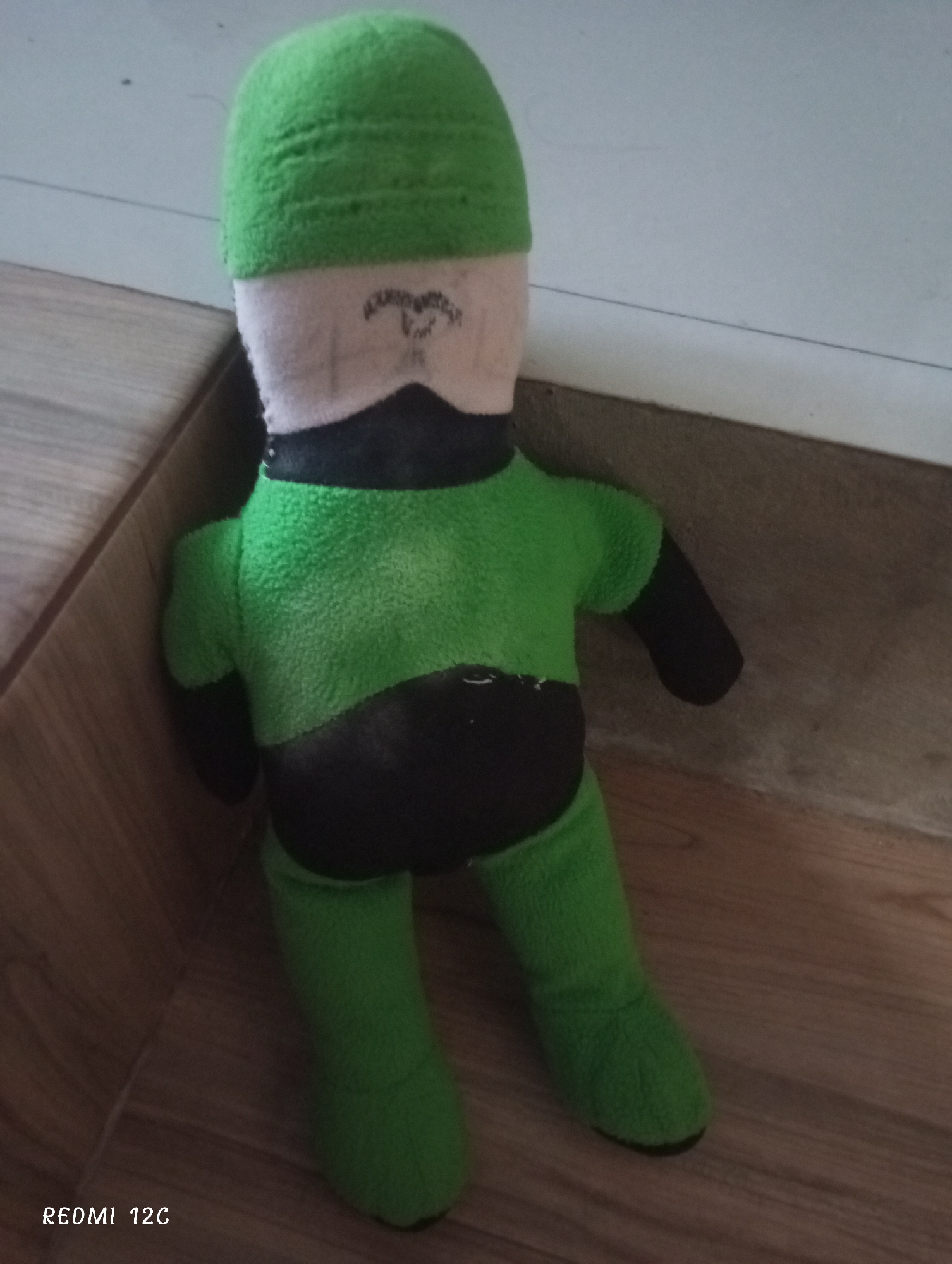 A stuffed, plush doll stands indoors in a corner formed by a light-colored wall and wood-paneled flooring or a low shelf. The doll has a green cap, light-colored face with a hand-drawn facial marking, a black neck area, green torso, black lower torso, black arms, and green legs. The camera is labeled "Redmi 12C" in the lower left. There's no indication of a specific location in Lhokseumawe, Indonesia.