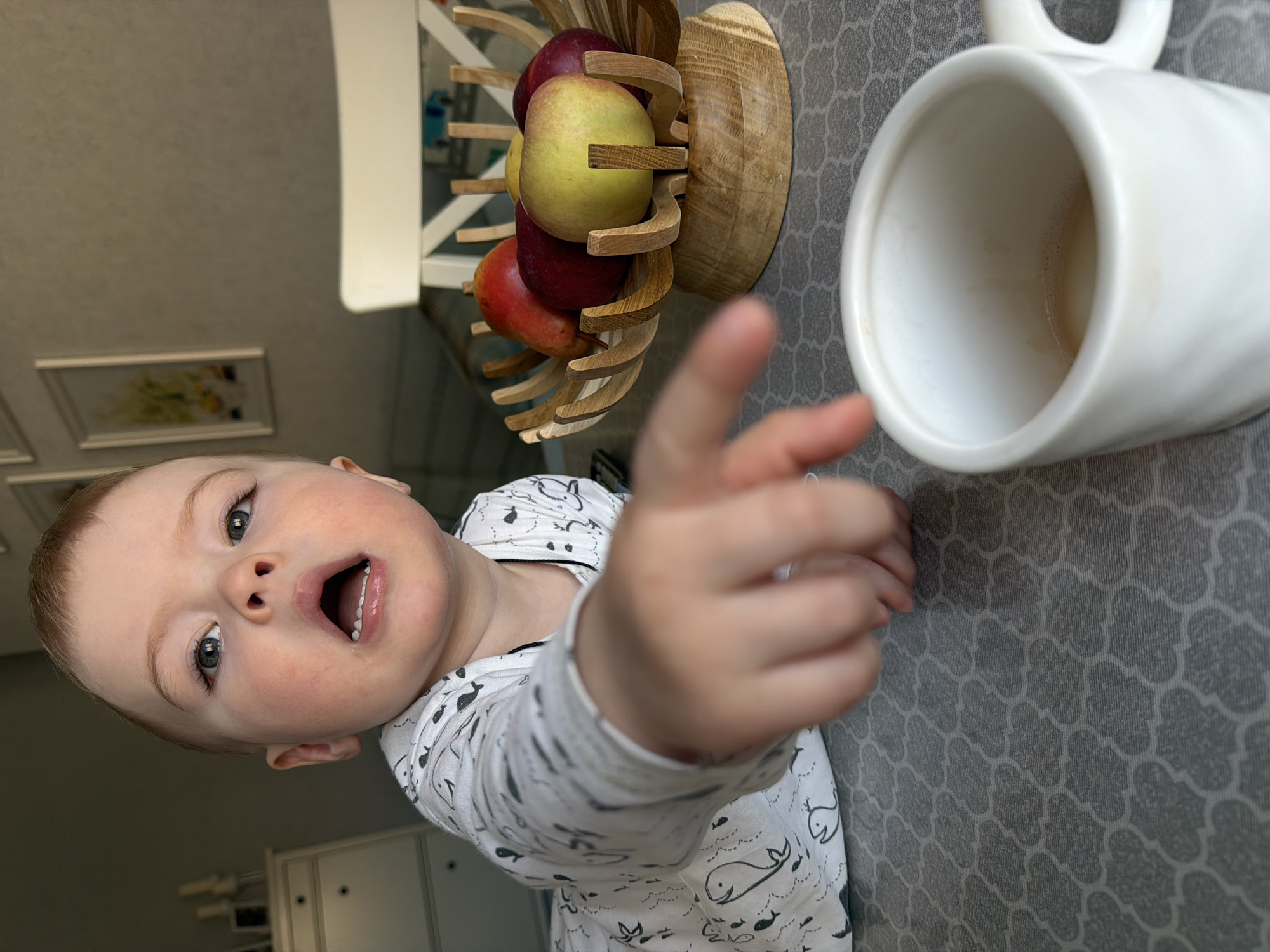 A young child, wearing a white pajama top with whale patterns, is pointing towards a white mug and a wooden fruit bowl with apples on a grey patterned surface. The photograph appears to be taken from a low angle, looking up at the child and the objects on the surface. The mug appears to have a small amount of liquid at the bottom. There is a white chair visible in the background and cabinets can be seen. The location is a room with common household items and based on the presence of the child and domestic objects, it could be a home or residence. The city of Votkinsk, Russia cannot be verified from the image.