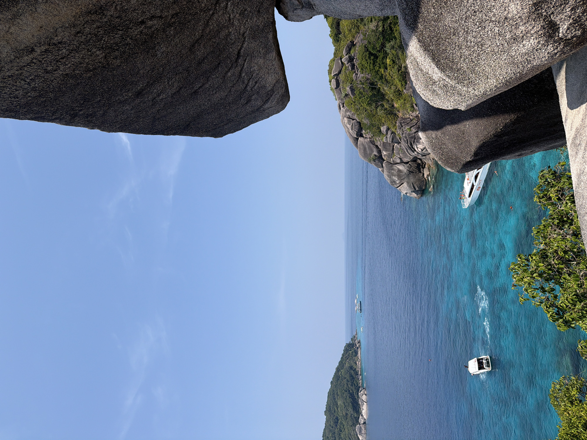 The scene depicts a tropical marine landscape within the Similan Islands Archipelago, located off the coast of Lam Kaen, Thailand. The perspective is from an elevated rocky vantage point.
The foreground on the right is dominated by large, dark grey, weathered granite boulders, one of which significantly overhangs the view, exhibiting a rough, granular texture.
Below, the clear, calm ocean water displays varying shades of blue, transitioning from deep indigo in the distance to vibrant turquoise closer to the islands. In the mid-ground, a white speedboat, labeled "ONCE" on its hull, is anchored in the turquoise water. Two individuals are visible in the water near this speedboat; one is equipped with an orange buoyancy aid. Further to the left, a second white boat is in motion, generating a visible white wake in the water. Several smaller, indistinct vessels are present on the distant horizon.
Two distinct islands or landmasses are visible across the water. The island on the far left is densely covered in green vegetation, with light grey boulders along its shore. Another island to the right is also vegetated and characterized by numerous large, smooth, dark grey granite boulders. The background features a clear, light blue sky with a few wispy white clouds.