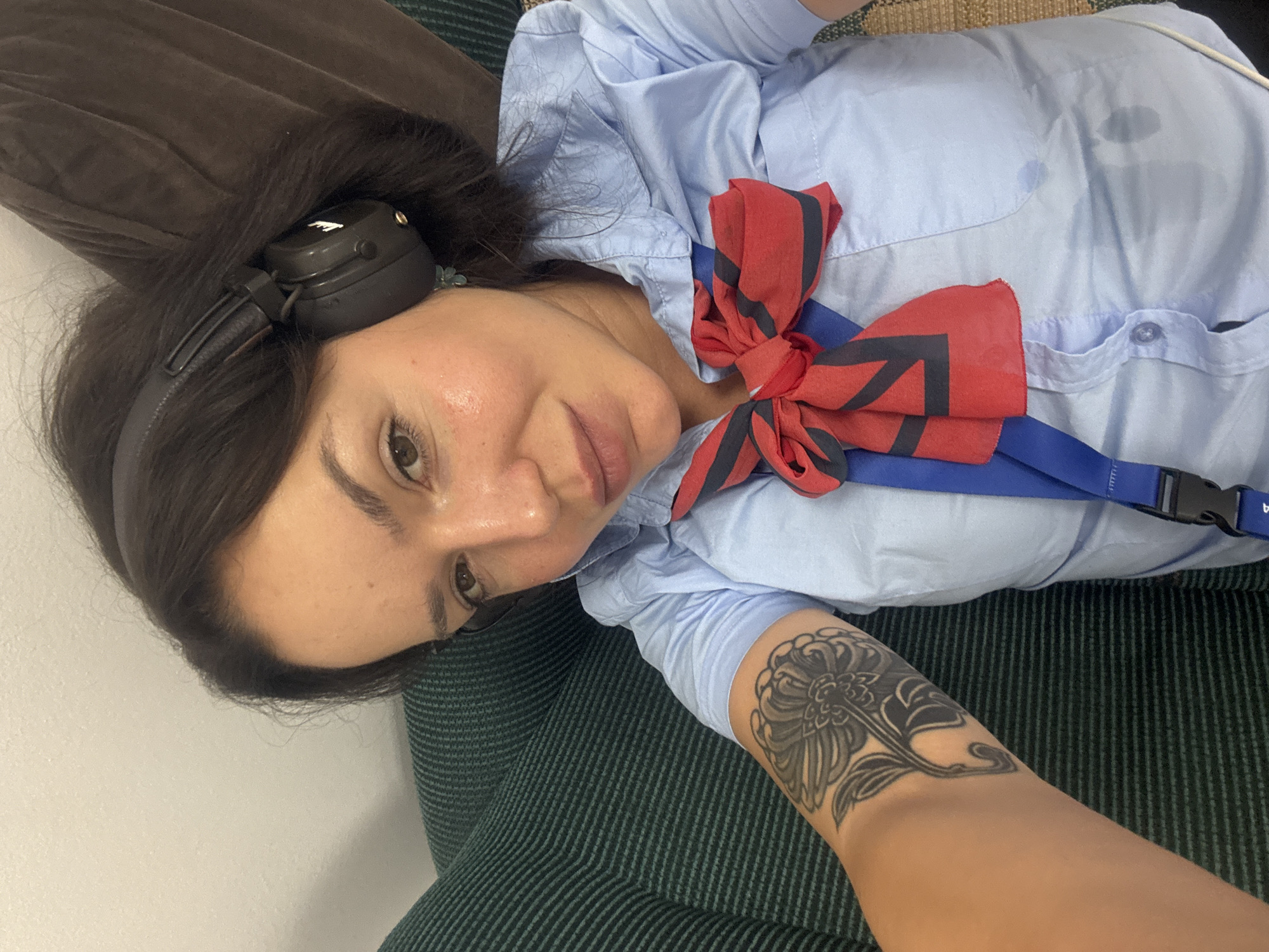 The image shows a woman lying on her side, wearing a light blue collared shirt and black headphones. She has dark hair and a tattoo on her left arm. A red and black striped bow is pinned to her shirt, along with a blue lanyard. The scene appears to be indoors, possibly on a couch or bed, with a dark green striped surface visible beneath her. The woman is looking towards the camera with a relaxed expression. There are no obvious time of day or weather cues, but the indoor setting suggests it could be any time. No visible text is present in the image. The context provided indicates the location is Burgas, Bulgaria.