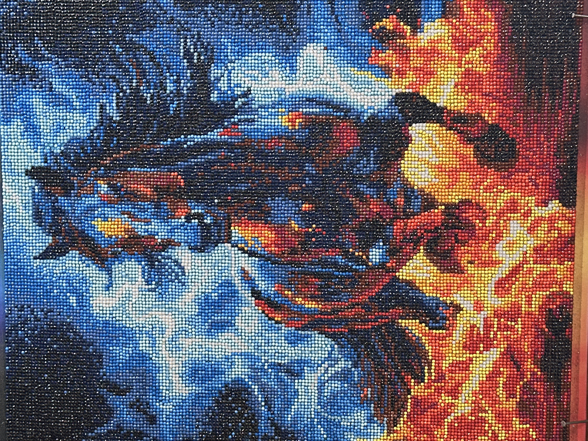 The image depicts a dragon made of small, colored beads. The dragon is composed primarily of blue, black, and red beads. The left side of the image features a cooler color palette with shades of blue and white, suggesting an icy or water-based environment. The right side is dominated by warmer colors, with shades of red, orange, and yellow, suggesting a fire-based environment. The dragon's body seems to be a mix of both elements, blending the cool and warm colors. The overall composition gives the impression of a duality or conflict between ice and fire elements. The location information is not conveyed or perceivable from the visual content alone.