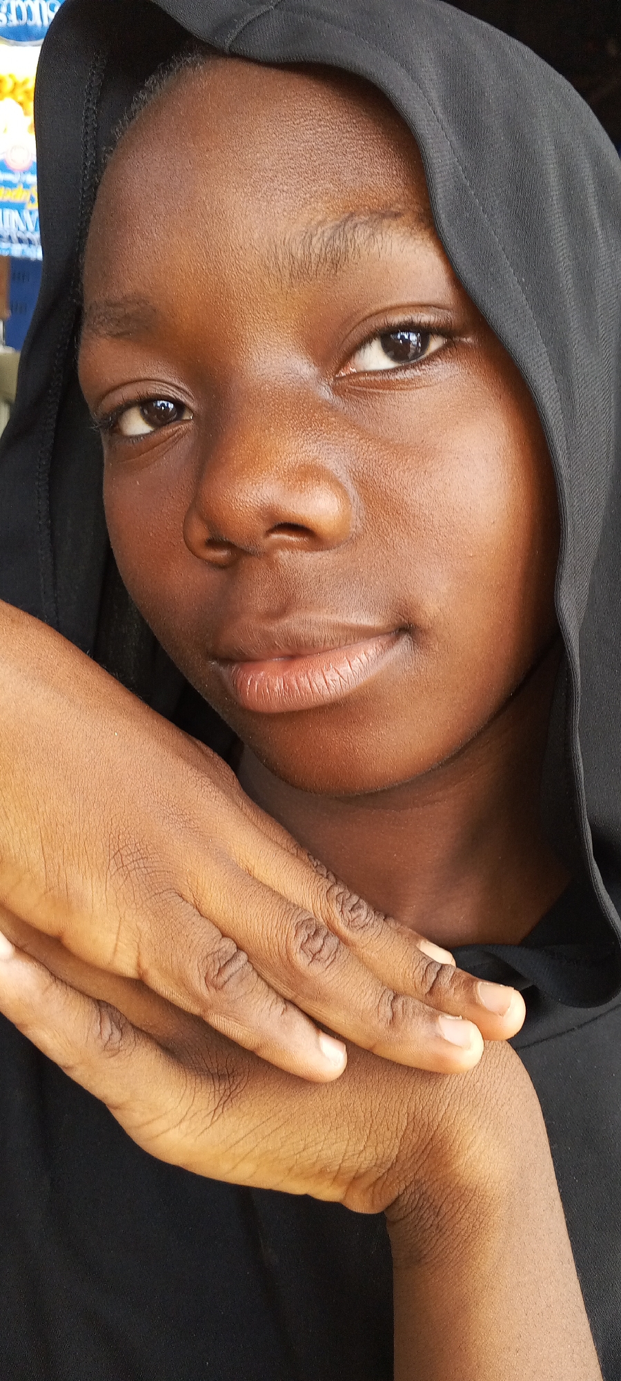 A close-up portrait captures a young individual, likely female, with a dark complexion, looking directly at the viewer. She wears a black hoodie or head covering that frames her face. Her expression is calm and gentle, featuring a subtle, closed-lip smile, and her dark eyes are bright and engaging. Her hands, also dark-skinned, are positioned in the foreground, appearing to be resting on her chest or shoulder, adding depth to the composition. The skin texture on both her face and hands is visible, showing natural detail.
The lighting is soft and even, suggesting the photograph was taken in an indoor setting or a well-shaded outdoor area during daylight hours. The background is softly blurred but reveals hints of a larger environment. Visible elements include fragmented blue, white, and yellow shapes, possibly from signs or advertisements, and partial text that includes "IT" and "NIVER" (potentially part of "UNIVERSITY" or a similar word). These background cues, combined with the location context of Lagos, Nigeria, suggest a public, possibly urban or commercial setting. The overall mood of the portrait is serene and intimate.