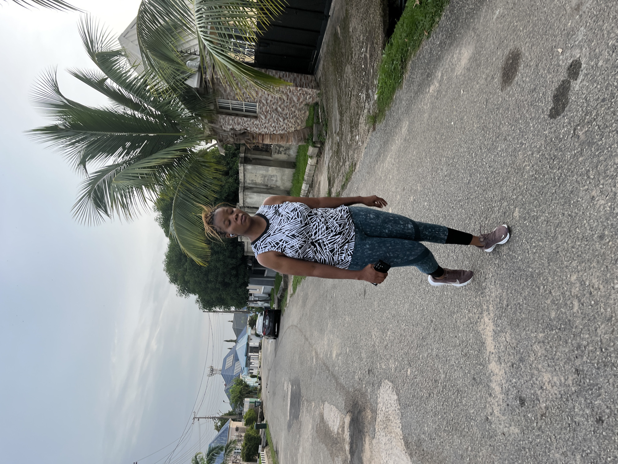 A woman with braided hair is walking on a paved street in Abuja, Nigeria. She is wearing a black and white patterned tank top, teal leggings with a pattern, and brown athletic shoes. She is holding a smartphone in her right hand. In the background, there are several houses with blue roofs, palm trees, and a dark-colored sedan parked on the side of the street. The sky is overcast.