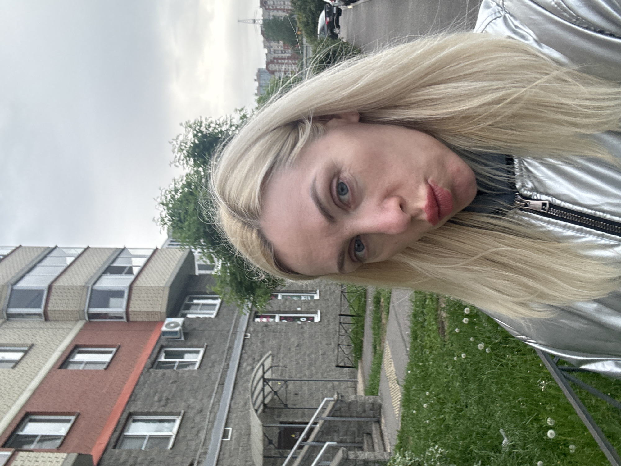 A medium shot captures a woman with light skin, blonde hair, and blue eyes in an urban environment of Unknown, Unknown. She is wearing what appears to be a silver jacket. The background features a building constructed of grey and red brick, and green vegetation. Above the building, a grey, overcast sky dominates the top of the frame. The photograph is taken at an angle, with the woman and building slightly tilted to the left.