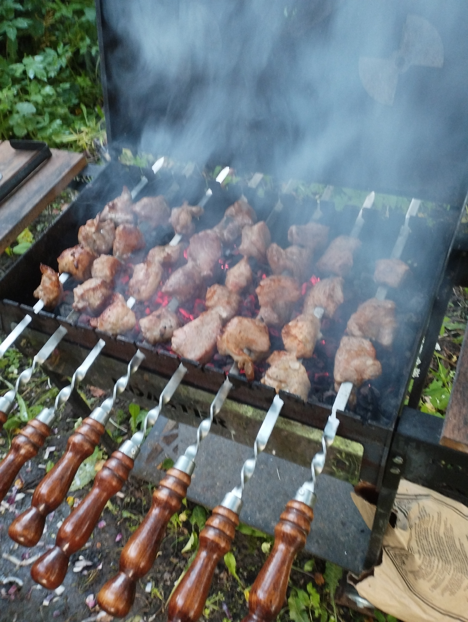 The image captures an outdoor cooking scene in the city of Ширяево (ильинский), Russia. A metal barbecue grill is filled with skewers of meat cooking over hot coals, producing a visible plume of smoke. The meat is cut into bite-sized pieces and appears to be browning as it cooks. Several skewers are visible in the foreground, each with a polished wooden handle and a twisted metal shaft. The grill itself is situated on the ground, surrounded by sparse green vegetation and fallen leaves, suggesting a natural or park-like setting. The background is blurred but shows more foliage, indicating the presence of trees or bushes. The overall impression is that of a casual outdoor meal being prepared.