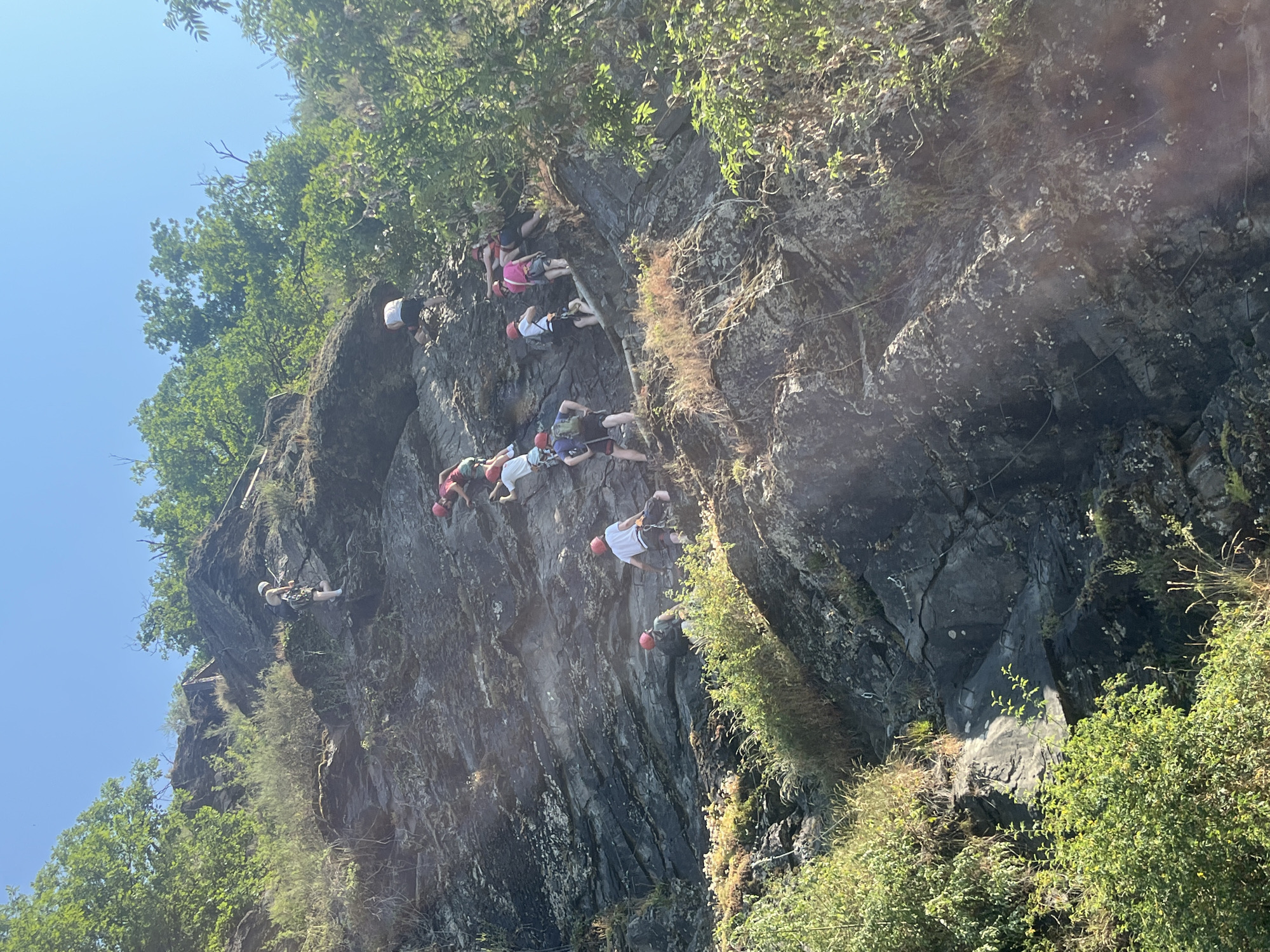 In an outdoor, rocky, mountainous environment, a group of people are rock climbing. The climbers, wearing helmets and harnesses, are ascending a steep, dark gray rock face. Green trees and vegetation are visible on the slopes and at the top of the rock face. The sky above is clear and blue. The scene suggests an activity that requires physical exertion and skill.