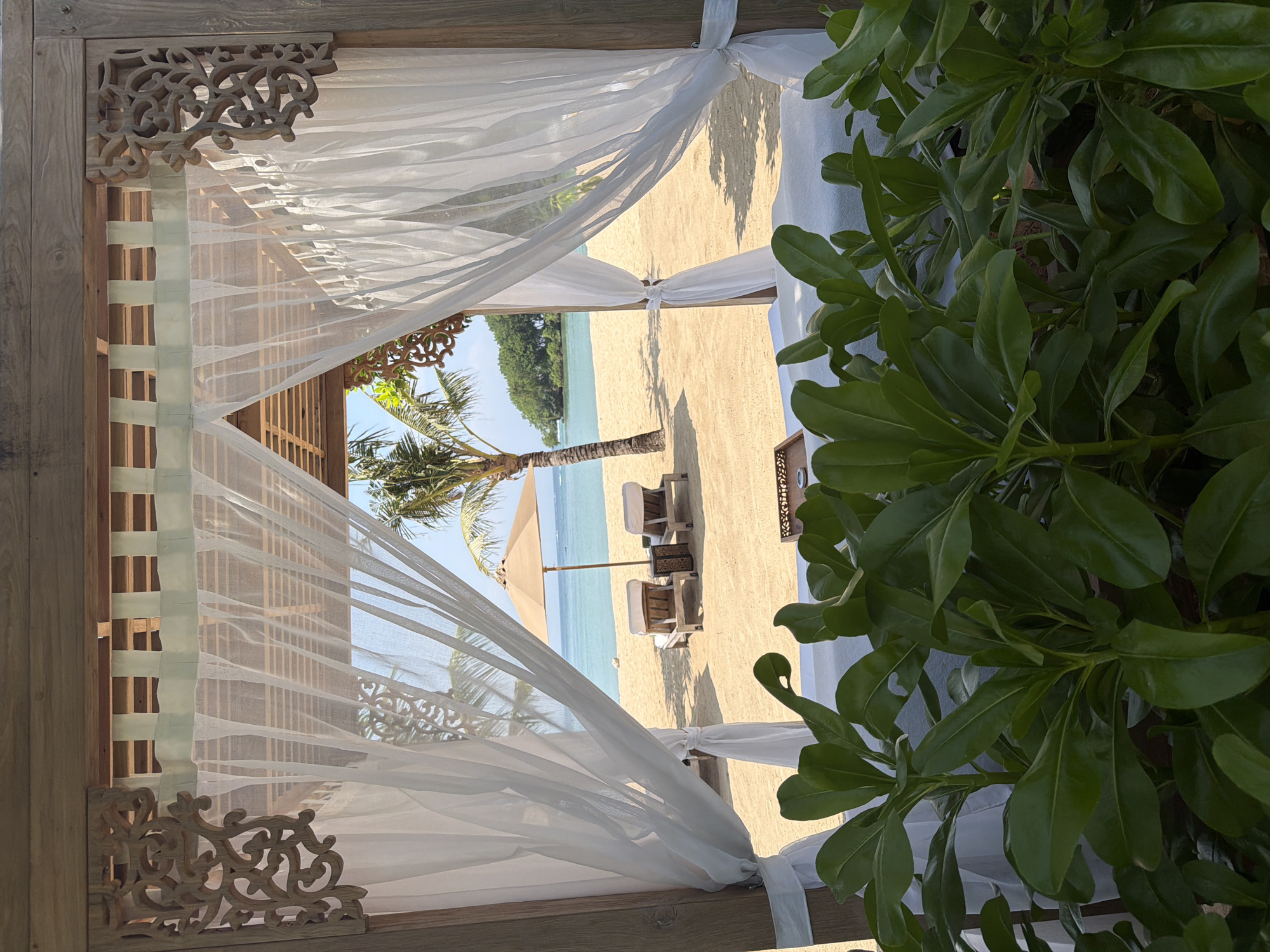 The image depicts a tropical beach scene in Nusa Dua, Indonesia, viewed from within a wooden cabana or gazebo. The foreground is dominated by lush green foliage with broad leaves. Framing the central view, a light-colored wooden structure features carved decorative elements in its upper corners and has sheer white curtains tied back to its posts. In the midground, a sandy beach is visible under clear skies. Two empty wooden lounge chairs with white cushions are positioned on the sand, facing the calm turquoise ocean. A large, open beige parasol stands between and slightly behind the lounge chairs. A single palm tree is present on the sand, casting a shadow to its right. In the background, the ocean extends to the horizon, with a small, heavily vegetated island visible on the right side of the water. No individuals are present in the scene.