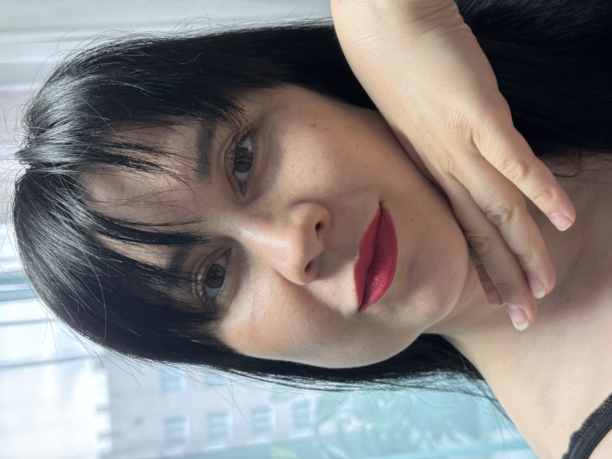 A woman with dark hair and red lipstick poses indoors. Her left hand is near her face. The background includes a window and a building. There is no indication of the location being Zav'yalovo, Russia.